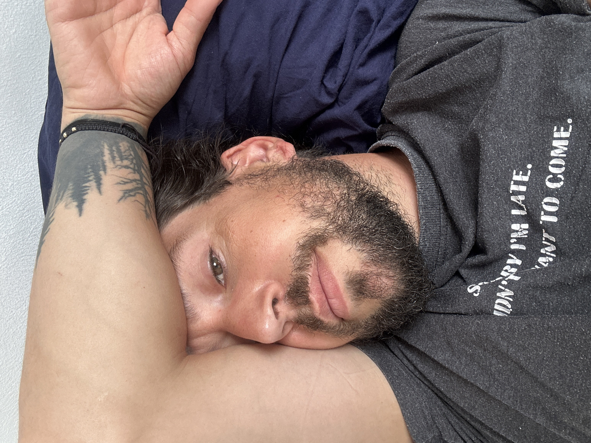 A man is lying on his side on a blue pillow and wearing a gray T-shirt with white text. His right arm is raised and bent with a bracelet on his wrist and a tattoo of trees on his arm. The man has brown hair and a beard. The background is a plain white wall. The location in the city of Nivelles, Belgium, is not discernible from the image.
