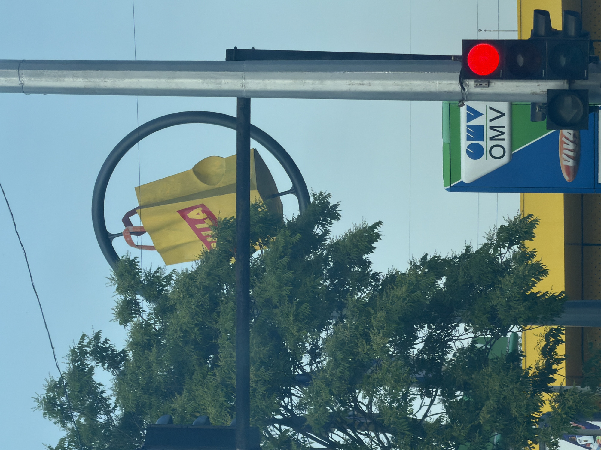 The image presents an outdoor urban scene in Shumen, Bulgaria, under a clear, light blue sky. A large, dark-colored circular frame or ring structure is prominently displayed in the upper center, partially obscured by a vibrant green deciduous tree on the left. Suspended within this ring is a three-dimensional sculpture of a yellow shopping bag with red handles. Partial red lettering "ULA" is visible on the bag's surface, suggesting a supermarket or retail brand like "KAUFLAND".
To the right, a thick, multi-faceted grey metal pole stands vertically, supporting a traffic light assembly. The top section of the traffic light displays a solid red signal. Below the main traffic light, a secondary traffic light with no illuminated signal is visible.
Further down and to the right of the tree, a blue and green sign with the white "OMV" logo is visible, indicating a fuel station. Below the OMV logo, a red oval sign with the word "VIVA" in white is present. Behind the OMV sign and the lower part of the tree, a structure with a bright yellow facade is discernible, likely a commercial building. Several dark overhead wires traverse the sky horizontally in the upper part of the frame. No individuals are visible in the scene.