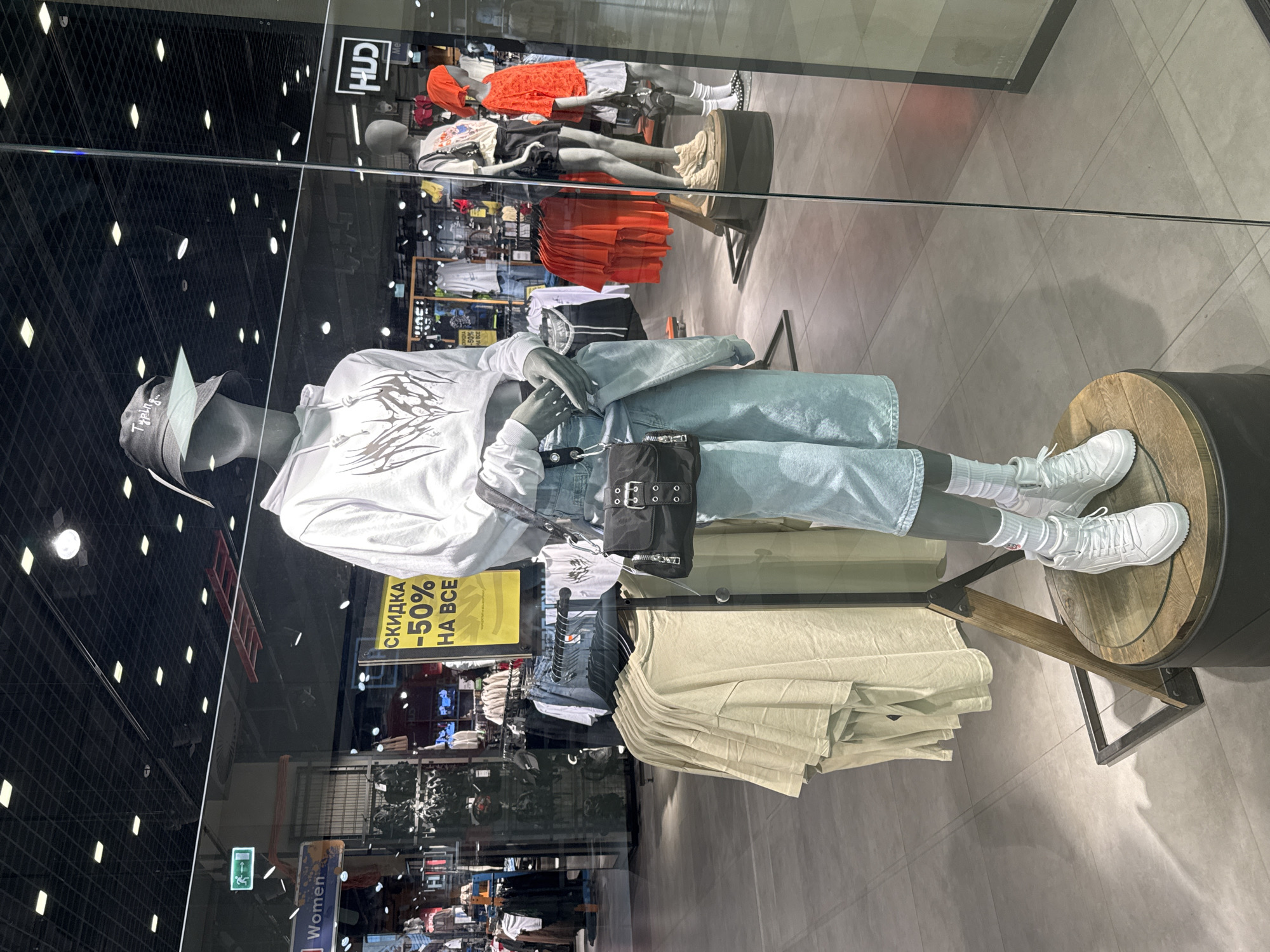 A photograph shows a department store window display at the "HUD" store in the city of Saint Petersburg, Russia. A mannequin is dressed in a white hoodie with an abstract design, light blue denim shorts, white socks, and white sneakers. It wears a bucket hat and a small black purse on its shoulder. The mannequin stands on a small, round wooden platform. Behind it, other mannequins display clothing, including orange tops and black shorts. A yellow sign partially visible advertises "СКИДКА -50% НА ВСЕ," which translates to "DISCOUNT -50% ON EVERYTHING". The store is well-lit with black grid lighting in the ceiling. Clothes racks stocked with various clothing items can be seen inside the store through the window's reflection. A red ladder and a green exit sign are visible in the window's reflection. The floor inside the store and the floor in front of the display are tiled.