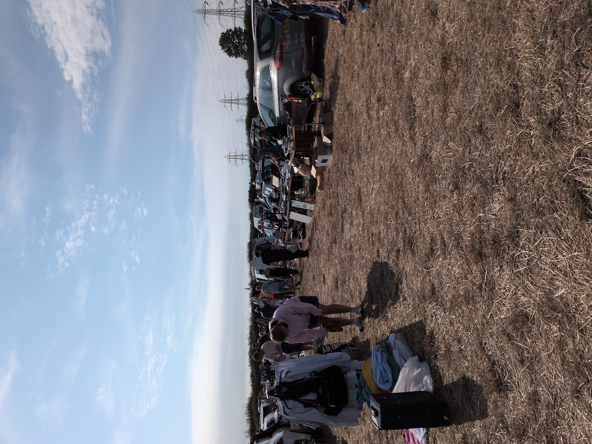 This is a full, eye-level, outdoor, daytime, vertical shot of a car boot sale, likely in South Staffordshire, United Kingdom. The ground is covered in dry, matted grass, indicative of a field or large open area. A cloudy, blue sky with scattered white clouds is visible in the upper portion of the image. In the background, several parked cars and vans are present, along with rows of stalls set up beside them. Electrical pylons and trees line the horizon.
In the foreground, a black suitcase and a stack of folded blankets and clothing are laid out on the ground. To the left, a clothing rack holds various garments, including a white shirt and a black handbag. A woman wearing a pink hooded sweatshirt and dark shorts is bent over, looking at her shoe. Other people are scattered throughout the scene, browsing the stalls or standing near their vehicles. The overall impression is one of a busy outdoor market on a bright, sunny day.