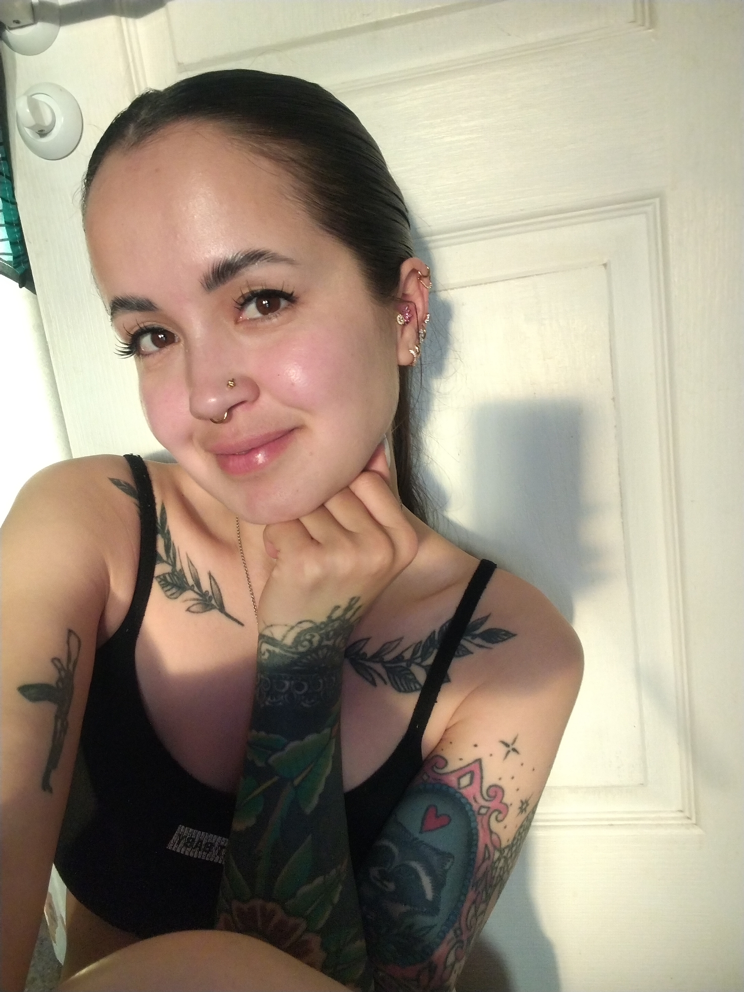 A woman is posing for a selfie indoors against a white door. She has dark hair pulled back, light skin, and several tattoos on her arms and chest. She is wearing a black tank top. She has multiple piercings in her ear and a nose ring. Her left hand is resting under her chin. The location is in an unidentified interior space; the city of Noviy, Russia is not depicted in the image.