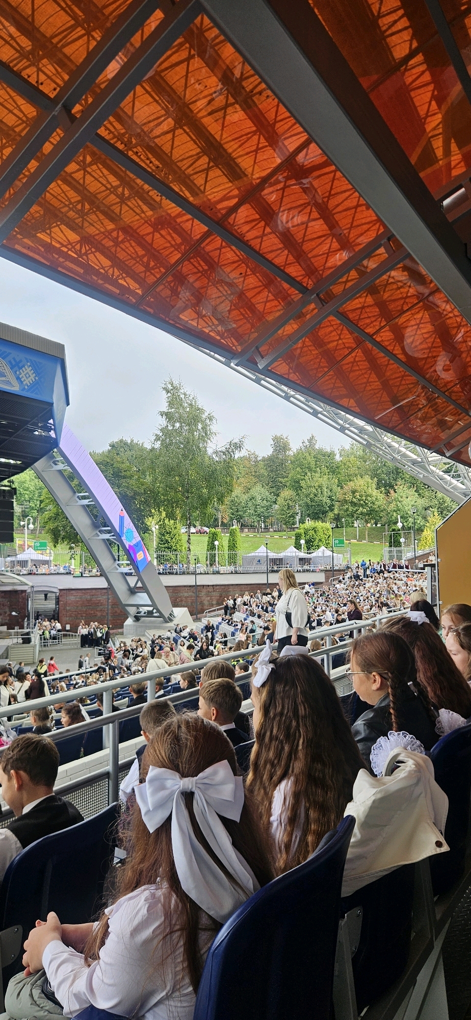 The image depicts an outdoor amphitheater filled with people, likely attending an event. The scene is viewed from the seating area, looking towards a stage and a backdrop of trees. The roof of the amphitheater is a prominent orange translucent material supported by a metal structure. In the foreground, several children with large white bows in their hair are visible, suggesting a school event or celebration. They are seated in dark blue chairs. In the background, a large crowd of people are seated, facing a stage area where a few individuals are standing. There is a large screen or digital display visible on a slanted structure. The overall atmosphere appears to be festive and communal. The location is identified as the Vitebsk Summer Amphitheater in Vitebsk, Belarus, a venue renowned for hosting the annual "Slavianski Bazaar in Vitebsk" festival.