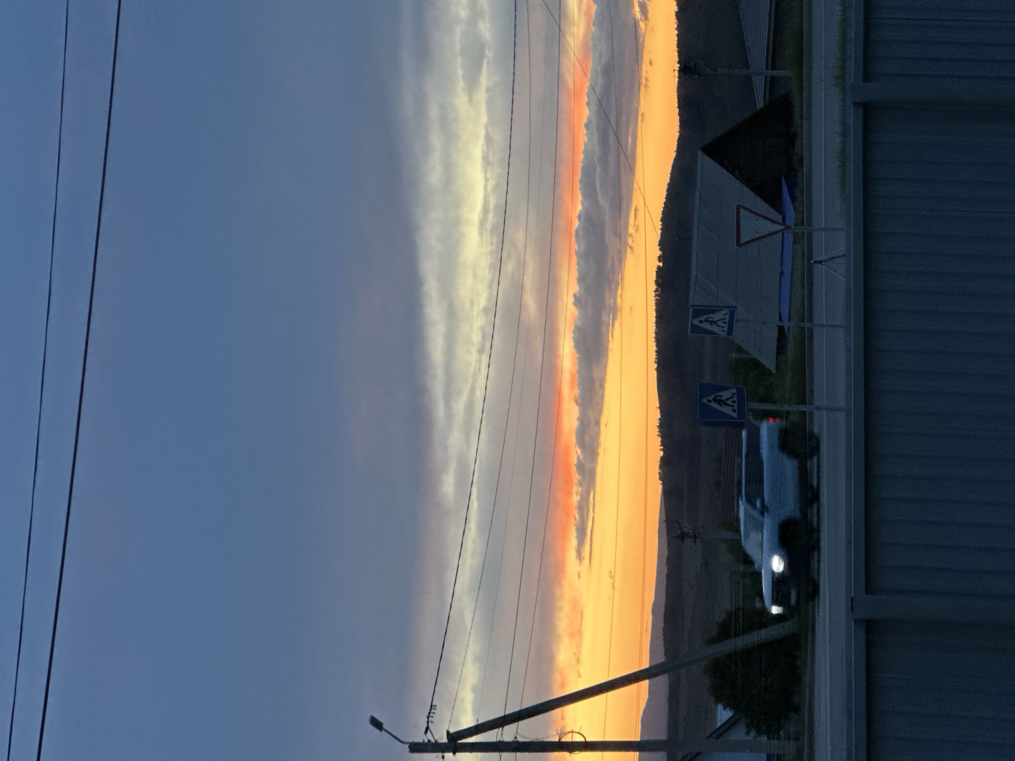 The image shows a landscape in Uchaly, Russia at dusk. The sky is a mix of blue and orange hues from the setting sun. There are power lines running across the upper part of the frame. In the right side of the image, there is a road with signs marking a pedestrian crossing, and a white SUV is driving on the road. A gray corrugated metal fence runs along the right edge of the frame. Behind the road, there is a dark hill or mountain range. There is a small building visible behind the pedestrian crossing sign.