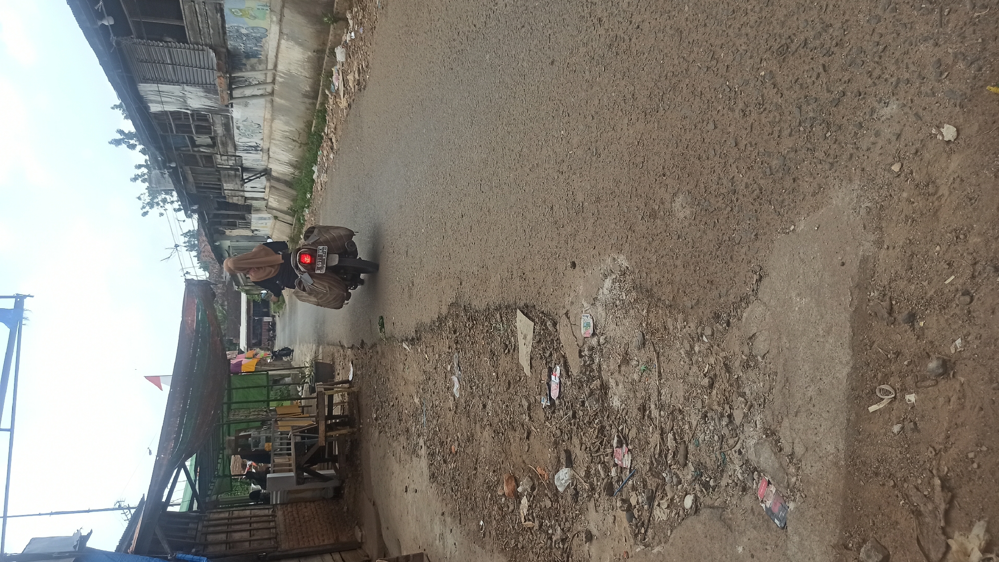 An outdoor, eye-level, vertical, medium shot shows a person on a scooter driving down a road in the city of Tuba, Indonesia. The person is wearing a tan hijab and is riding a scooter with two bags strapped to the back. The road is paved and cracked, with debris on the side. To the left of the road are some buildings made of corrugated metal and wood, with an awning covering a small area. To the right of the road are more buildings, also made of corrugated metal and wood. The sky is overcast.