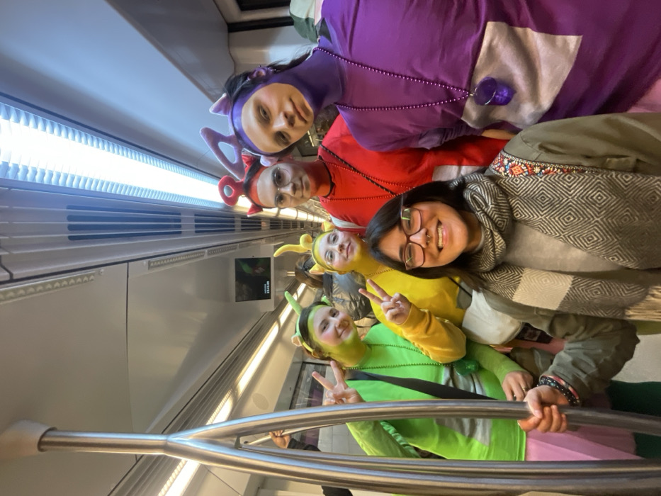 Six individuals are present inside a modern subway car in Barcelona, Spain. The car features white walls and ceiling, with continuous strip lighting and an overhead digital screen displaying "13:41" and what appears to be route information. A polished silver vertical grab pole is prominent in the foreground.
In the center foreground, a smiling individual with dark hair, glasses, and a nose ring holds the grab pole with their right hand. They wear a dark green jacket and a grey and white patterned scarf. To their left, two individuals are dressed in Teletubby-themed costumes: one in bright green with green face paint and antennae, holding up two peace signs; the other in bright yellow with yellow face paint and antennae, holding up one peace sign. Both wear matching beaded necklaces. To the right of the foreground individual, two more individuals wear Teletubby-themed costumes: one in red with red facial markings and a red triangular antenna, also wearing glasses and a red beaded necklace; and another in purple with purple face paint, purple triangular antennae, and a purple beaded necklace with a large purple pendant. These four costumed individuals are positioned closely behind the foreground person, looking towards the camera. A sixth person's arm is partially visible on the far left, grasping a handle in the background.