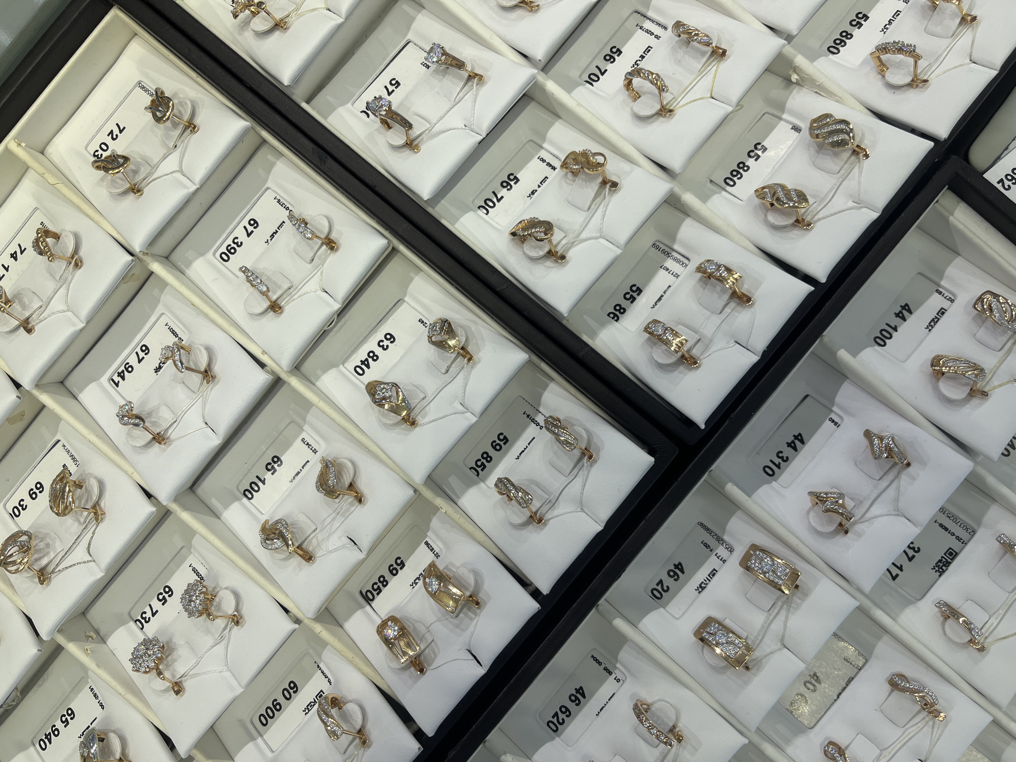 The image shows a display of gold earrings in individual compartments within a jewelry store, possibly located in Chaikovsky, Russia. Each compartment contains a pair of gold earrings presented on a small white display card, with a tag indicating a price or item number. The earrings vary in design and style, some featuring embedded stones or intricate metalwork. The compartments are arranged in rows and columns within a dark-colored tray, creating a grid-like pattern. The tags and small size of the jewelry make it clear these are individual, high-value items presented for retail sale.