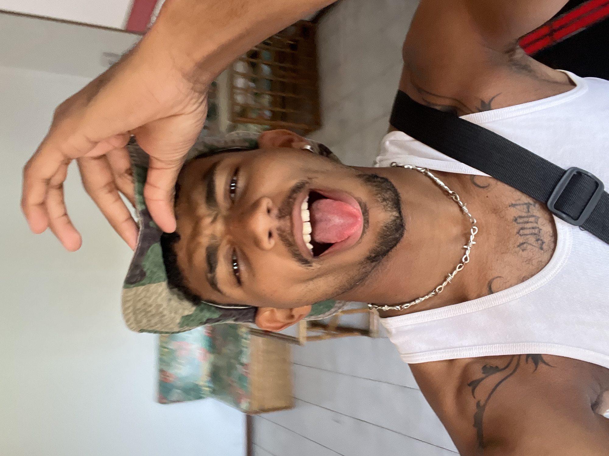 A young man with dark skin and a beard/stubble is captured in a close-up, rotated portrait, likely a selfie. He appears to be in Porlamar, Venezuela. He is wearing a white tank top and a camouflage-patterned cap. His mouth is wide open, with his tongue extended, and his eyes are wide, conveying a playful, energetic, or excited expression. He has a silver chain necklace, resembling barbed wire, and an earring.
Several tattoos are visible on his upper body. On his right shoulder/chest area, the numbers "2004" are tattooed, along with some indistinct script or design below it. A tribal or vine-like design is tattooed on the left side of his neck/upper shoulder. A black strap, possibly from a bag, crosses his chest over his white tank top. His left hand is raised towards his head, near his cap, possibly in a gesture or adjusting his hat.
The background is softly blurred, suggesting an indoor or sheltered outdoor setting with ambient daylight. Elements of the background include light-colored walls or surfaces, what appears to be wooden furniture or shelving, and a patterned fabric, possibly a cushion or curtain.