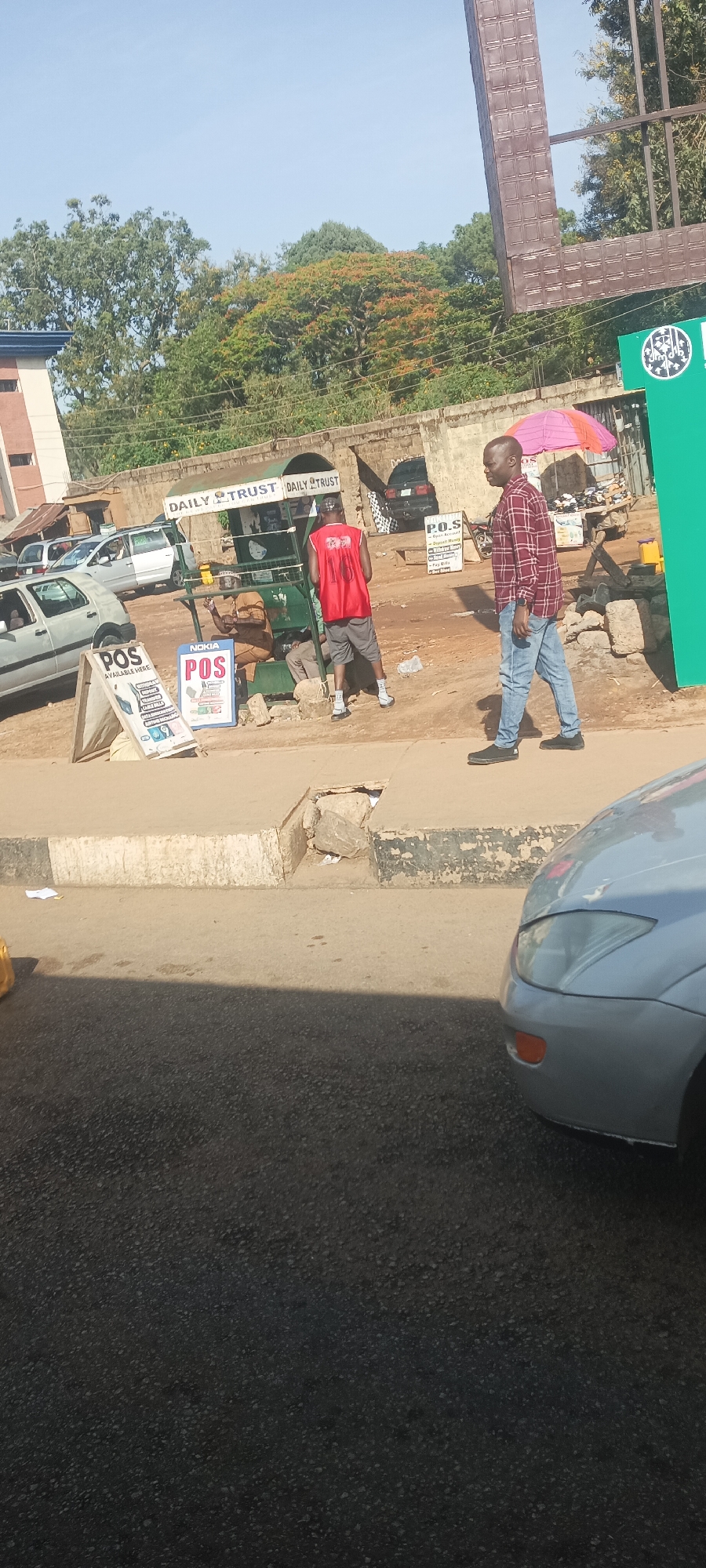 This image captures a vibrant street scene in Jos, Nigeria, during daytime under a clear blue sky. The setting includes a paved road, a concrete sidewalk, and a dirt area where commercial activities are taking place.
In the mid-ground, a green metal kiosk, prominently branded with "DAILY TRUST," stands as a central point of activity. A man in a light brown traditional outfit is seated inside the kiosk. Standing with his back to the camera near the kiosk is another man wearing a red sleeveless shirt and grey shorts. An A-frame sign directly in front of the kiosk advertises "P.O.S AVAILABLE HERE" and "NOKIA POS," indicating that mobile money and financial transaction services are offered. Further back, another small stand with a pink and orange striped umbrella also displays a "P.O.S" sign.
Walking across the mid-ground from right to left is a man dressed in a red and black checkered long-sleeve shirt and blue jeans. In the background, several cars are parked along the side of the road, and a light grey car is partially visible in the lower right foreground. A low brick wall and various buildings line the background, topped by dense green foliage and trees, some exhibiting reddish-orange leaves. The bright sun casts clear shadows, highlighting the time of day. The scene depicts typical daily life and commercial exchange in an urban Nigerian environment.
