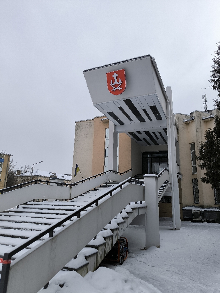 The image depicts the exterior of a building in Vinnytsia, Ukraine, under a cloudy winter sky. The building, identified by the Coat of Arms of Vinnytsia (a red shield with a silver anchor and two crossed sabers/swords) prominently displayed on an elevated canopy above its main entrance, features a light-colored facade, possibly beige or cream. A Ukrainian flag (blue and yellow) is visible on the building's left side.
Access to the main entrance is via a wide, concrete external staircase with black metal handrails. Both the stairs and the surrounding ground are covered in snow, with deeper snow accumulations at the base of the stairs. Beneath a section of the staircase, an orange portable generator is visible, connected by a white cable. To the right of the entrance, several external air conditioning units are mounted on the building's facade. A dark, evergreen tree is partially visible on the far right. The overall scene is devoid of people.