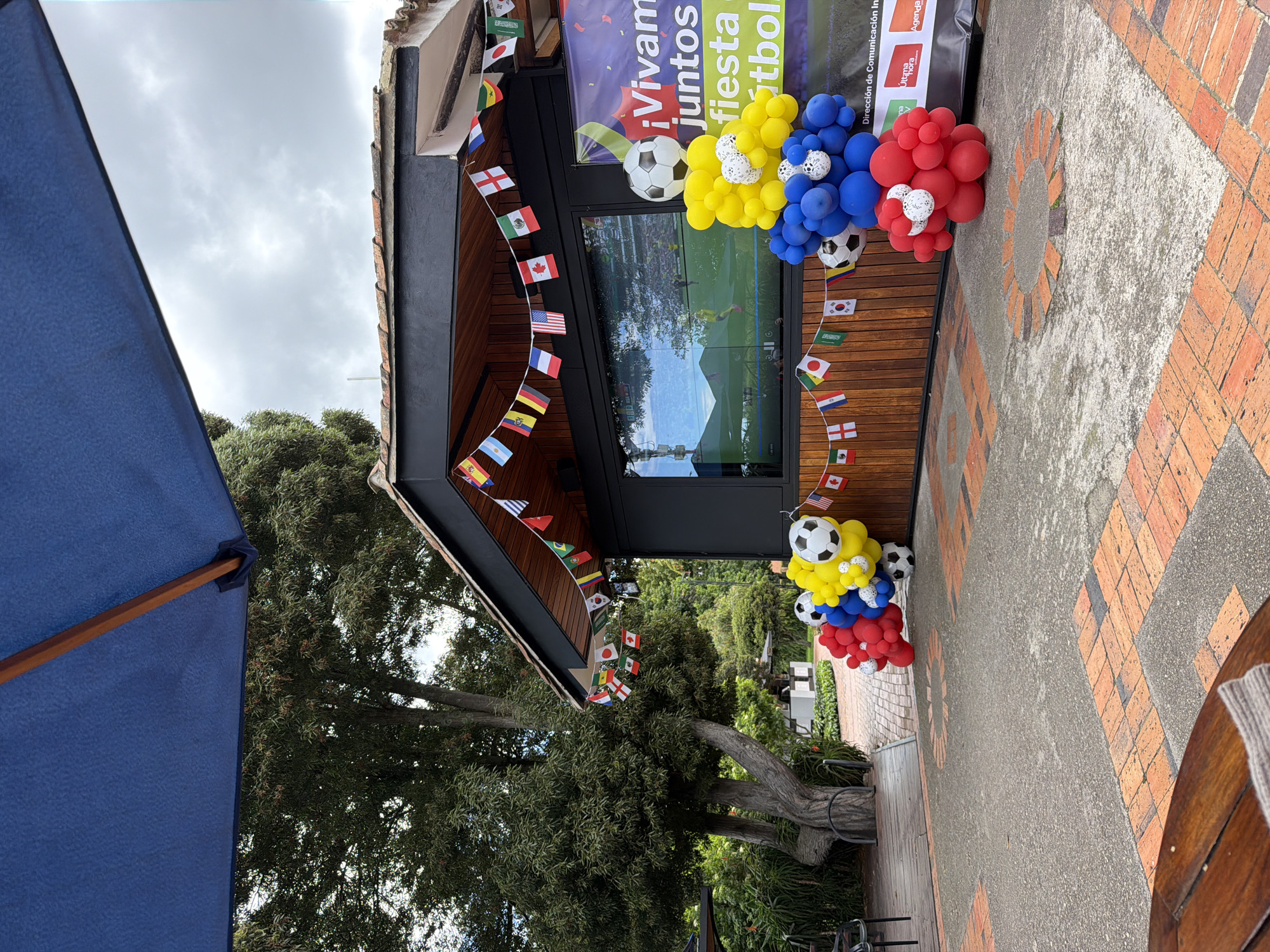 The image depicts an outdoor scene in Bojacá, Colombia, likely a commercial establishment decorated for a soccer event. A large blue umbrella provides shade, partially obscuring the upper left portion of the frame. Below the umbrella, a building with a dark-colored facade and a tiled roof is visible. A large window on the building's facade displays a soccer match being broadcast on a screen.
The building is adorned with a string of small international flags, including those of Spain, Argentina, France, the United States, Canada, England, Ghana, and Japan, among others. In front of the building, a vibrant balloon arrangement in red, blue, and yellow colors, interspersed with white balloons resembling soccer balls, stands prominently. Additional soccer ball-shaped balloons are placed around the arrangement.
To the right of the building, a large banner with the text "¡Vivamos juntos la fiesta del fútbol!" (Let's live the football party together!) is displayed. The banner also contains logos for "Última Hora" and "Agenda". A small Canadian flag is affixed to the building near the right edge of the frame.
The ground is paved with a combination of concrete and reddish-brown brickwork arranged in circular patterns. Lush green trees and foliage surround the area, suggesting a natural or garden-like setting. A wooden table and chair are partially visible in the foreground, indicating a seating area for patrons. The sky is overcast with clouds.