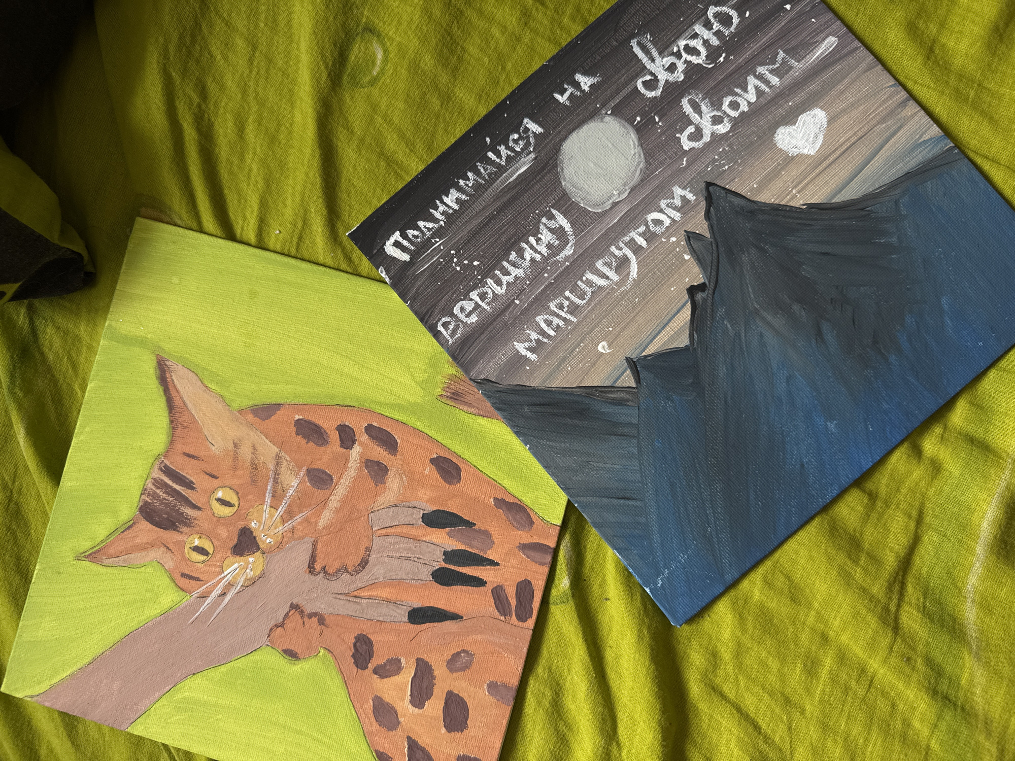 The image shows two square canvases on a green, textured surface. One canvas features a painting of a cat with a spotted coat clinging to a branch, set against a light green background. The other canvas depicts a mountain landscape at night with a moon, and white text in Cyrillic script. The paintings are situated side-by-side with the mountain canvas positioned at an angle to the cat canvas. The text on the mountain painting translates to "Climb to the top by your route".
There is no information regarding the location in the city of Izhevsk, Russia.