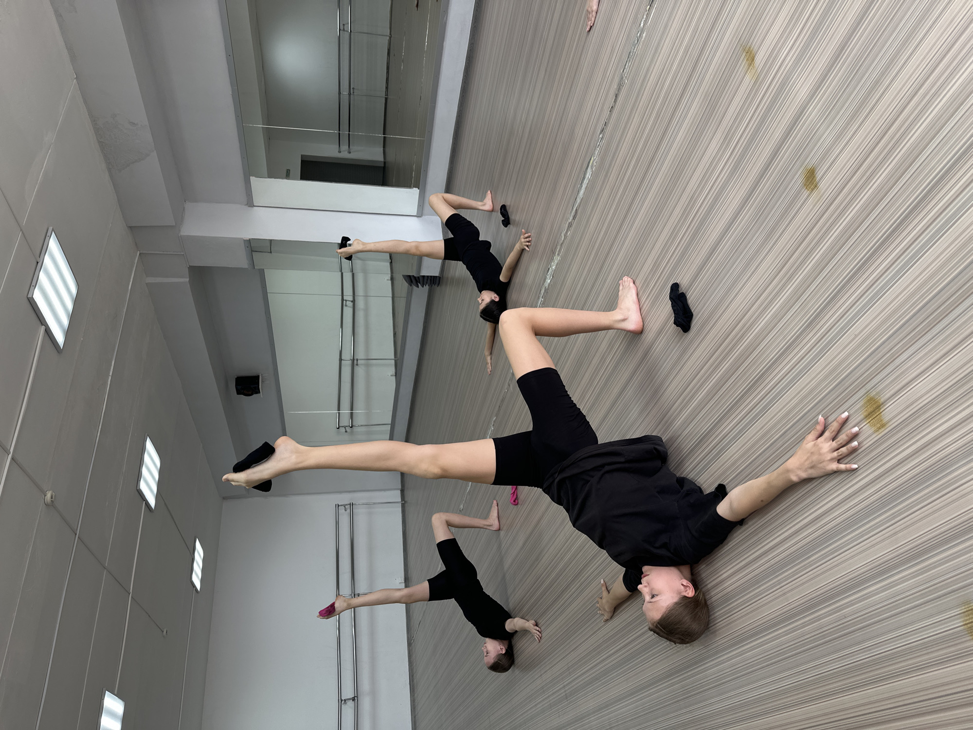 The image shows three individuals, likely dancers, in a dance studio. The studio has a wooden floor and mirrors on the wall. The individuals are performing a floor exercise, with their legs extended in different directions. They are wearing black clothing. A pair of socks is on the floor. The location is a dance studio in Votkinsk, Russia.