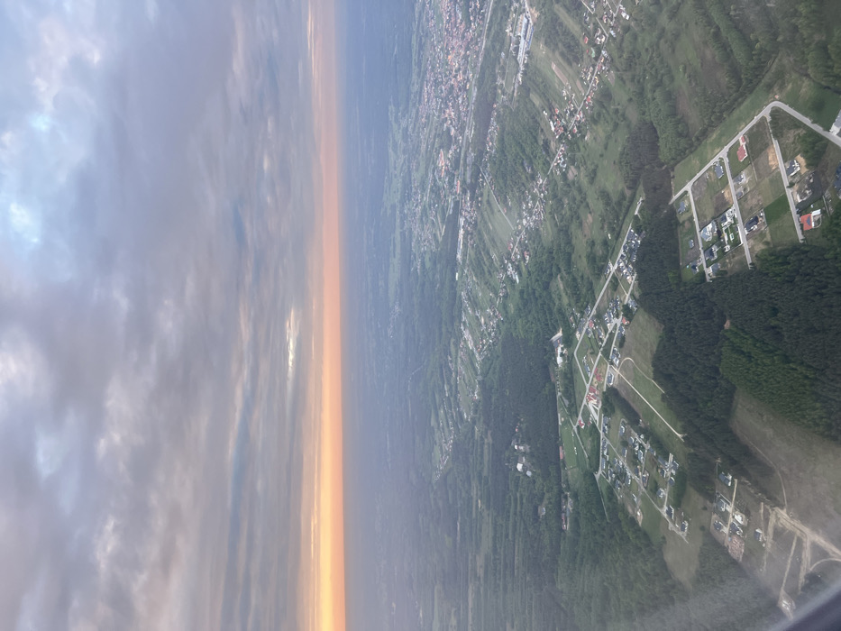 An aerial view of a suburban area in Jasionka, Poland (latitude: 50.109, longitude: 22.025) at sunset. The sky is a mixture of pink, orange, and gray clouds. The land is covered with a patchwork of green fields, forests, and houses. The image is taken from an aircraft, likely a passenger plane. The perspective is looking down at the scene from a high angle, creating a sense of distance. The image shows a suburban neighborhood with houses scattered across the landscape. Some houses are surrounded by trees, while others are in open fields. A long stretch of road bisects the neighborhood, and there are hints of a network of smaller roads. There are also patches of woods and fields, creating a variety of land cover types. There are no visible people in the image.