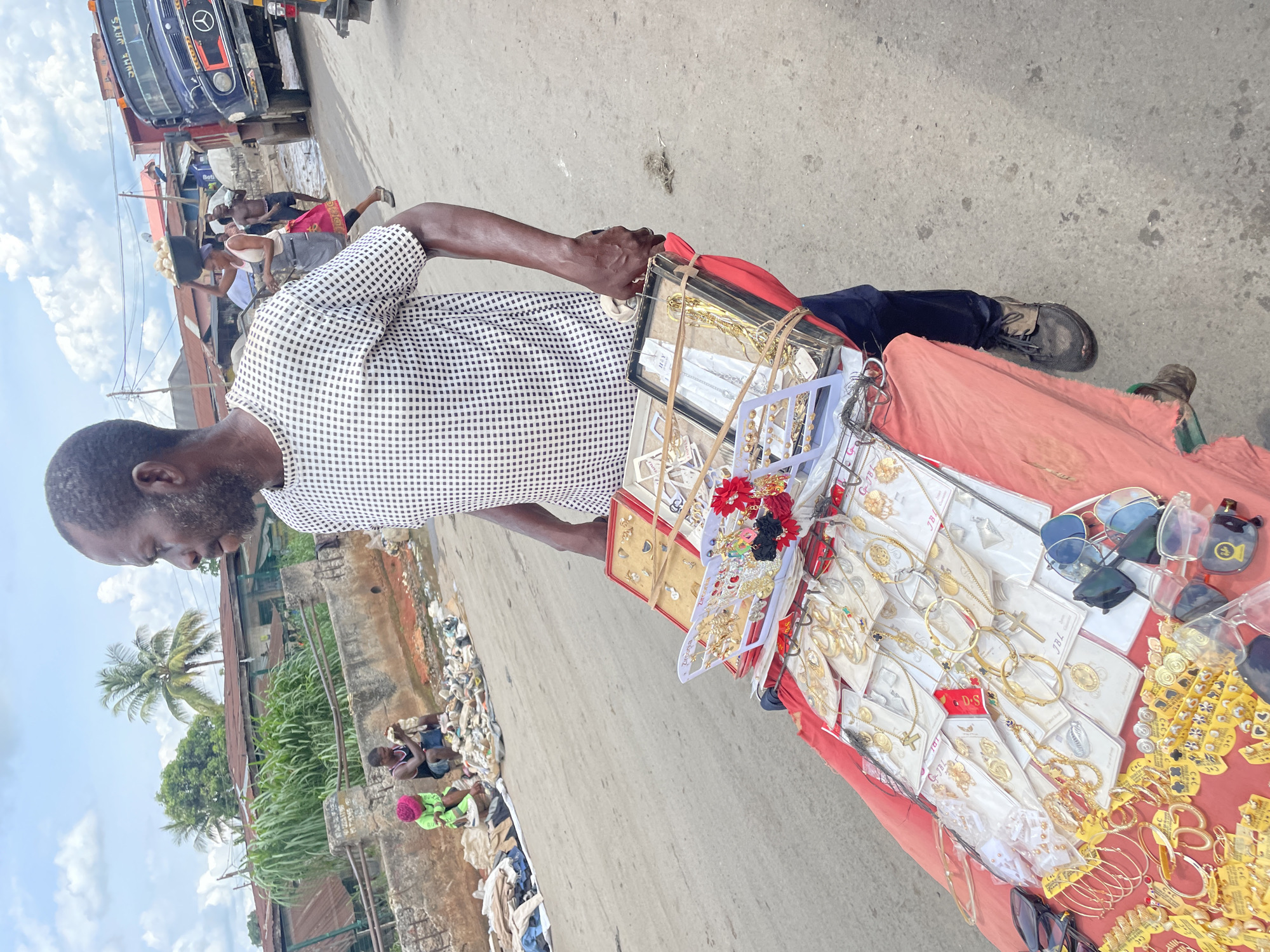 The image captures a vibrant street scene, likely in an open-air market or roadside commercial area, under bright daylight with a partly cloudy sky.
The central subject is a dark-skinned man with a beard, wearing a white and black checkered short-sleeved shirt and dark trousers. He stands on a paved surface, holding onto a mobile display cart covered with a red fabric. He appears to be a street vendor, showcasing a wide array of costume jewelry and accessories. The cart is laden with gold-colored necklaces, earrings (including hoops, crosses, and studs), bangles, and rings, many displayed on white cards in clear plastic packaging. Several pairs of sunglasses, including aviator and clear-framed styles, are also for sale. Hair accessories featuring red and black flowers are visible among the jewelry. Some jewelry items have "JBL" printed on their packaging.
In the mid-ground, a busy street stretches out behind the vendor. A large blue truck, possibly a Mercedes-Benz, with "SAW THIS" visible on its side, is parked or moving slowly. Various people are seen in the distance; one individual balances a bowl on their head. Further back, buildings with corrugated iron roofs and dense tropical vegetation, including palm trees, line the street. A partially visible blue sign reads "Betha".
To the right and further back from the vendor's cart, an open ditch or drain filled with a considerable amount of trash and debris (plastics, fabrics) is visible. Two individuals are seated or crouching near this area, one wearing a bright green top and pink head covering, suggesting they might be scavenging or working amidst the refuse.
The overall scene depicts a common sight in urban or semi-urban areas in some parts of the world, characterized by informal commerce and varied socio-economic activity.