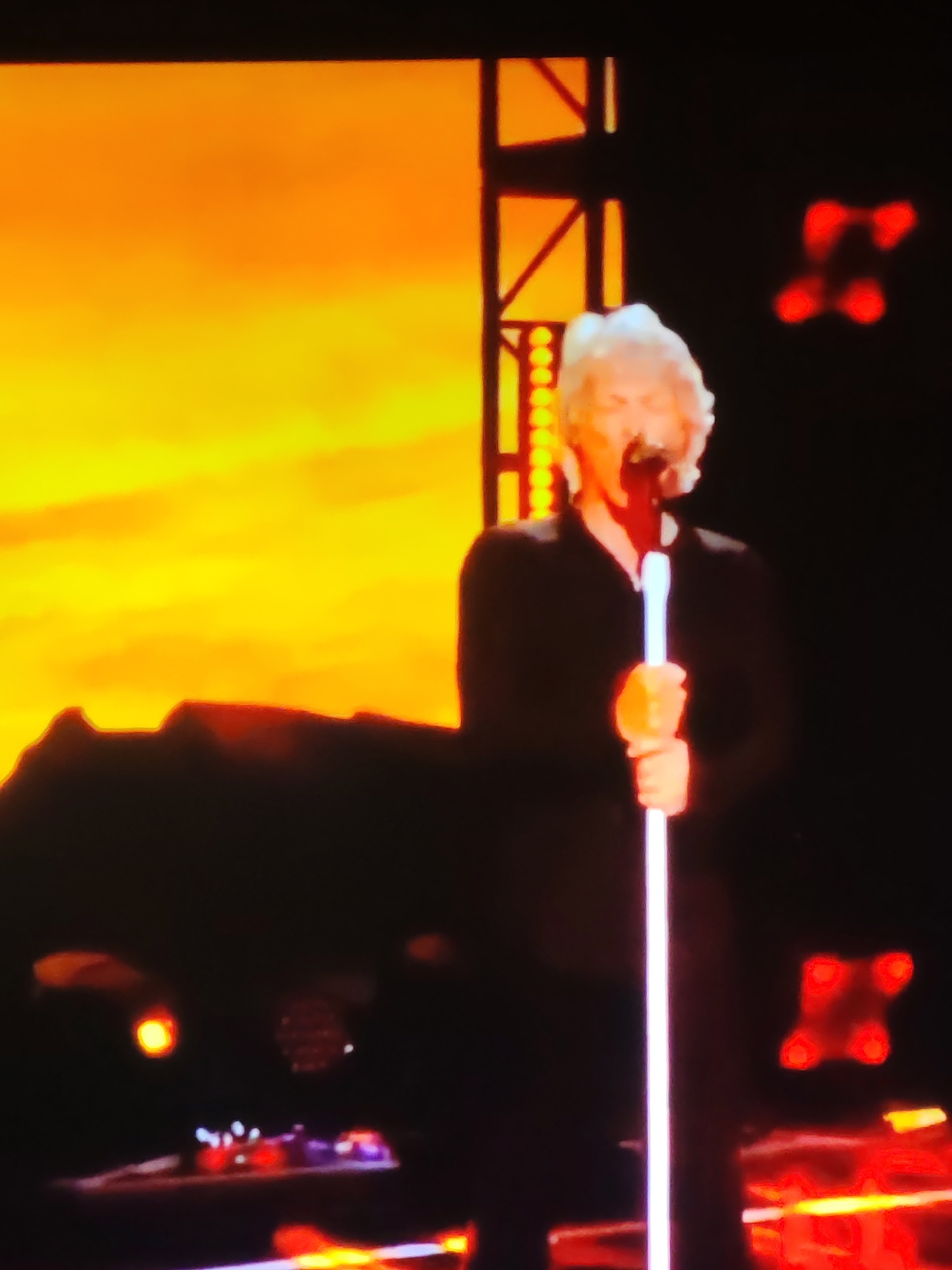 The image shows a man with white hair, wearing a black shirt and holding a microphone. He is singing into the microphone with his mouth open. Behind him, there is a large screen displaying a fiery orange and yellow background, with a dark silhouette of mountains or a landscape at the bottom. The stage is dark, with some lights visible in the background. There is no discernible information about the city of Vladimir, Russia, in the image.