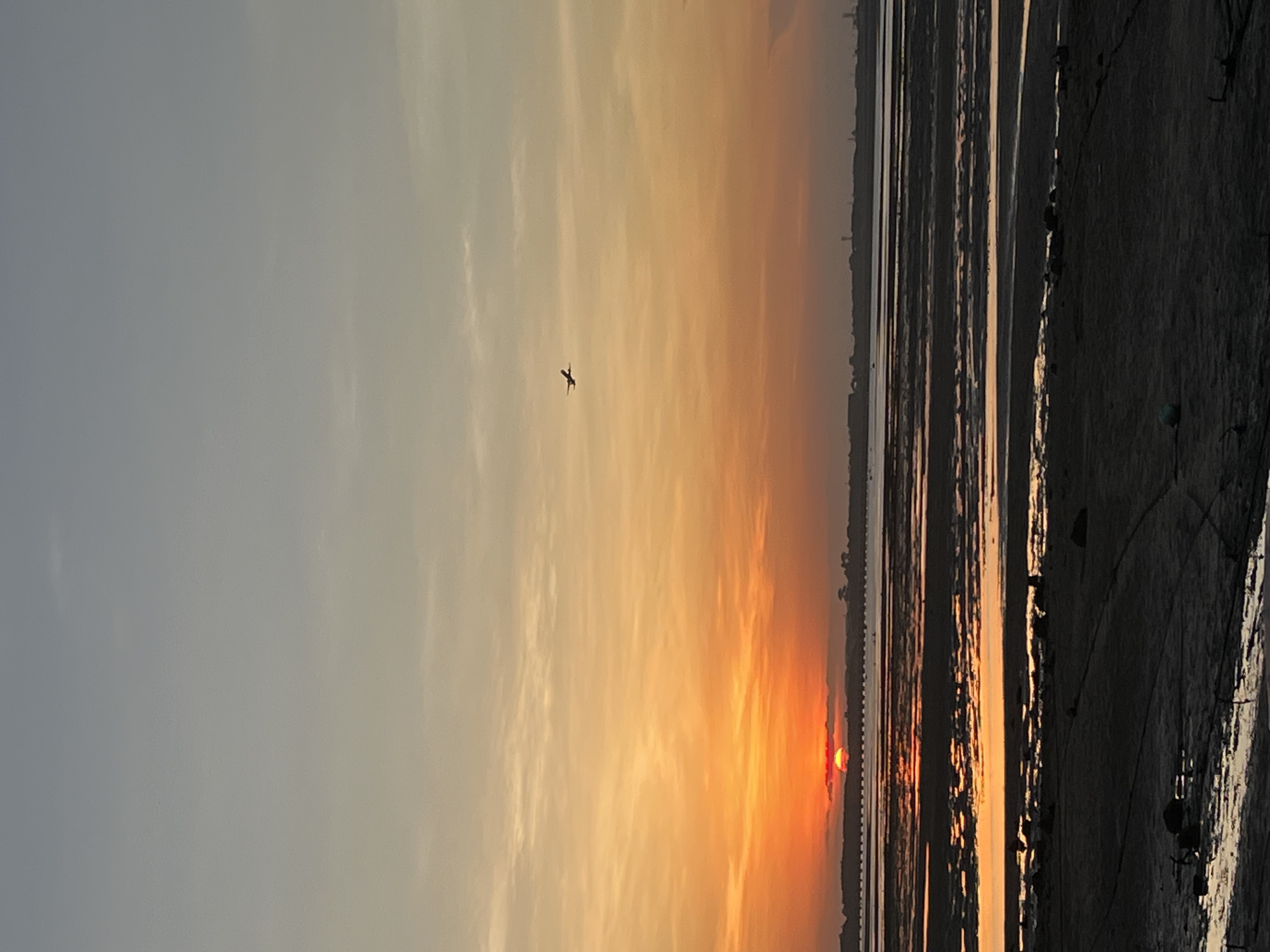 A horizontal photograph depicts a sunset scene, likely at a beach, in what may be Kuta Selatan, Indonesia. The sky transitions from a pale blue-gray to a vibrant orange near the horizon, where a red sun is setting, creating a stark contrast. A small bird is silhouetted against the sky. The foreground consists of textured, dark-colored sand or mudflats with grooves, possibly reflecting the sky. Vertical lines, seemingly from receding water, divide the land. In the distance, what could be a thin strip of land or a structure is visible on the horizon.