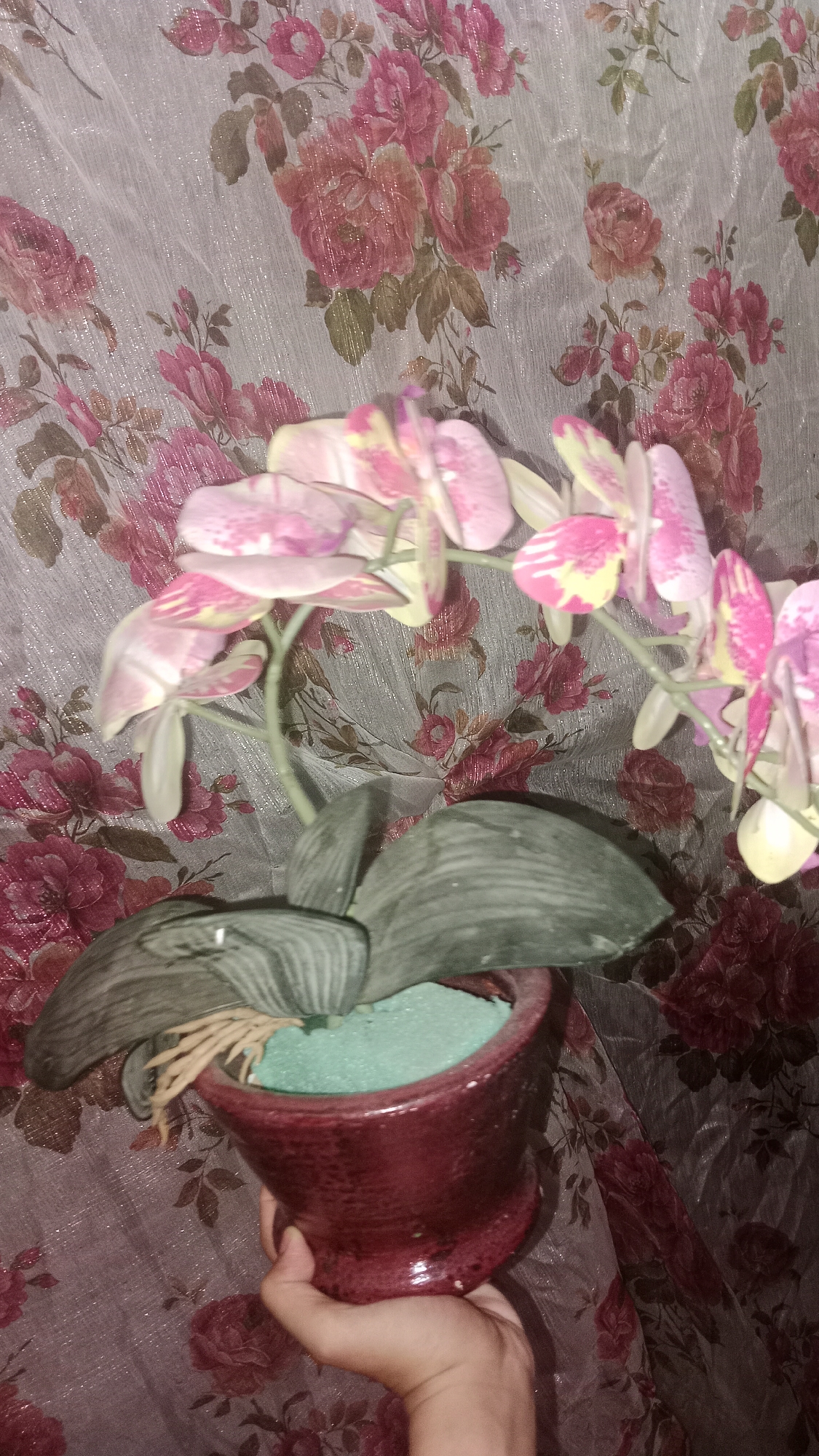 In Wono Sari, Indonesia, a hand is holding a dark red, glossy ceramic pot containing a faux orchid plant. The orchid features pink and yellow flowers with green leaves. The pot is filled with a light green, porous material. Behind the plant, a sheer fabric curtain with a pattern of pink and red flowers is visible. The lighting in the image is dim, casting shadows.