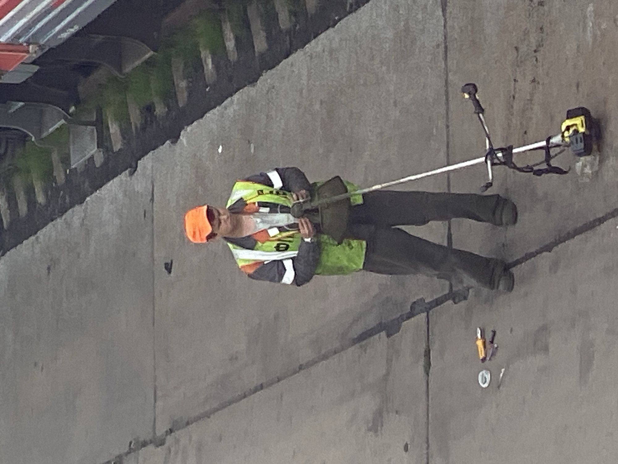 A worker wearing an orange cap, a reflective vest over a gray shirt, and dark pants stands on a concrete surface near train tracks. He is holding a string trimmer. In front of him on the ground are a roll of white tape and a small yellow and black tool. A portion of a red train car is visible to the right of the frame. The scene appears to be outdoors in an industrial or transportation setting, possibly a train yard or station platform.