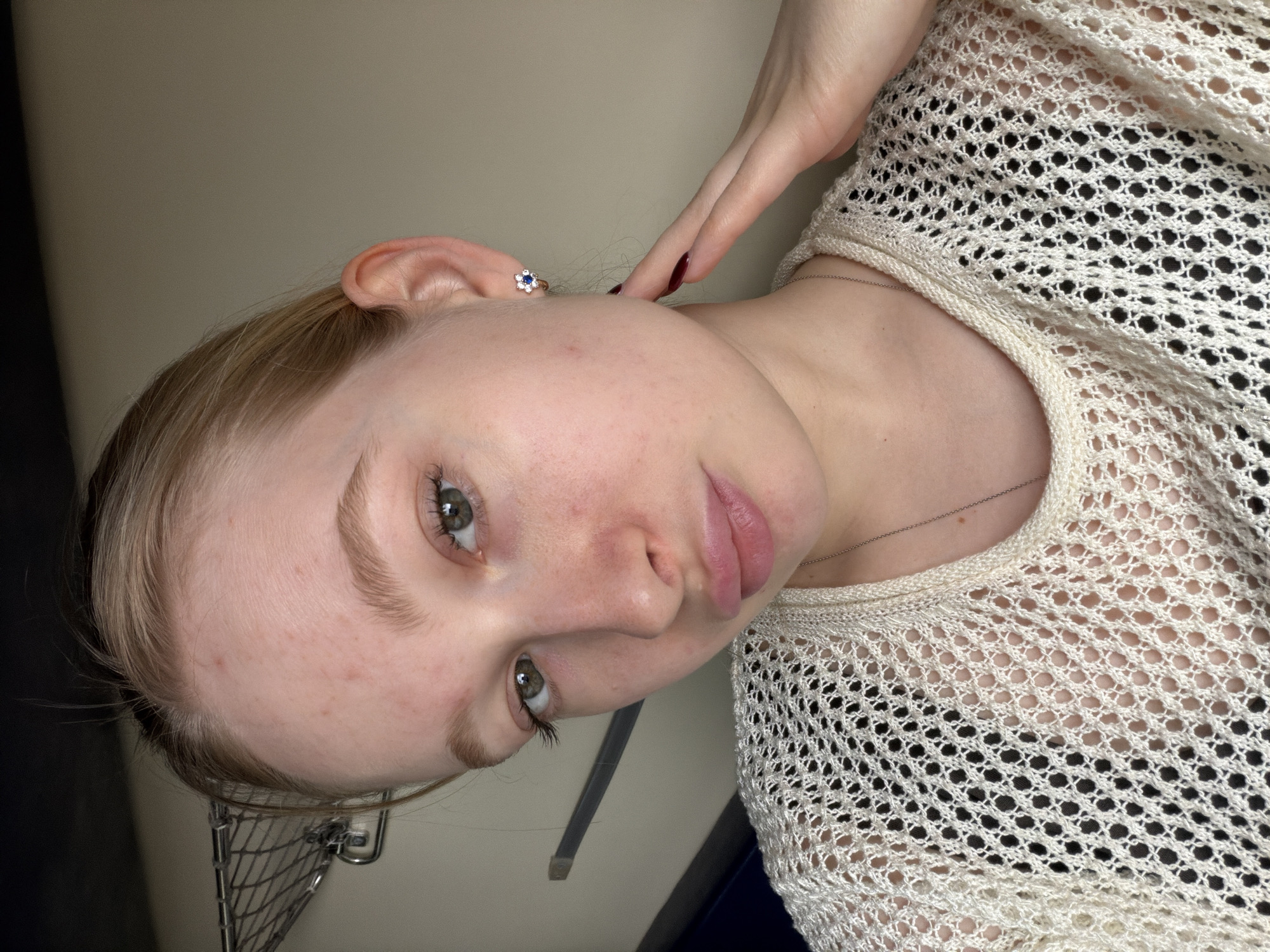 A close-up, portrait-style photograph shows a fair-skinned person with light blonde hair, appearing to be female, wearing a white, open-weave top. She has light eyebrows and visible eyelashes, with hazel eyes. The skin on her face shows some redness and possible minor blemishes. A small, floral-shaped earring is visible in her left ear. Her left hand touches her neck. A silver necklace can be seen near her collarbone. The background is a plain, light neutral color. A small, wire rack is visible in the bottom left corner of the image. There are no indicators this image was taken in Колышлей, Russia.