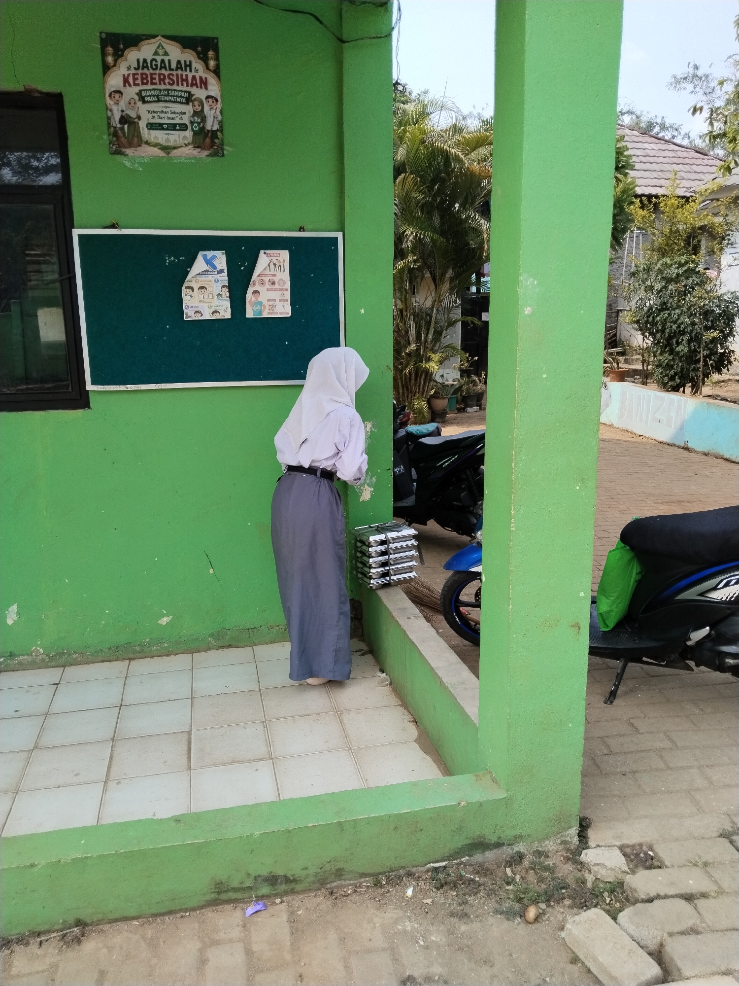 In Serang, Indonesia, a student wearing a white hijab and a grey uniform stands with their back to the camera, holding a stack of rectangular objects. The student is positioned in a corner with bright green walls and a tiled floor. A dark green bulletin board with posters is mounted on the wall behind the student. To the right, a blue and black scooter is parked, and further in the background, a tiled walkway and vegetation are visible. The scene appears to be an outdoor area of a school or public building.