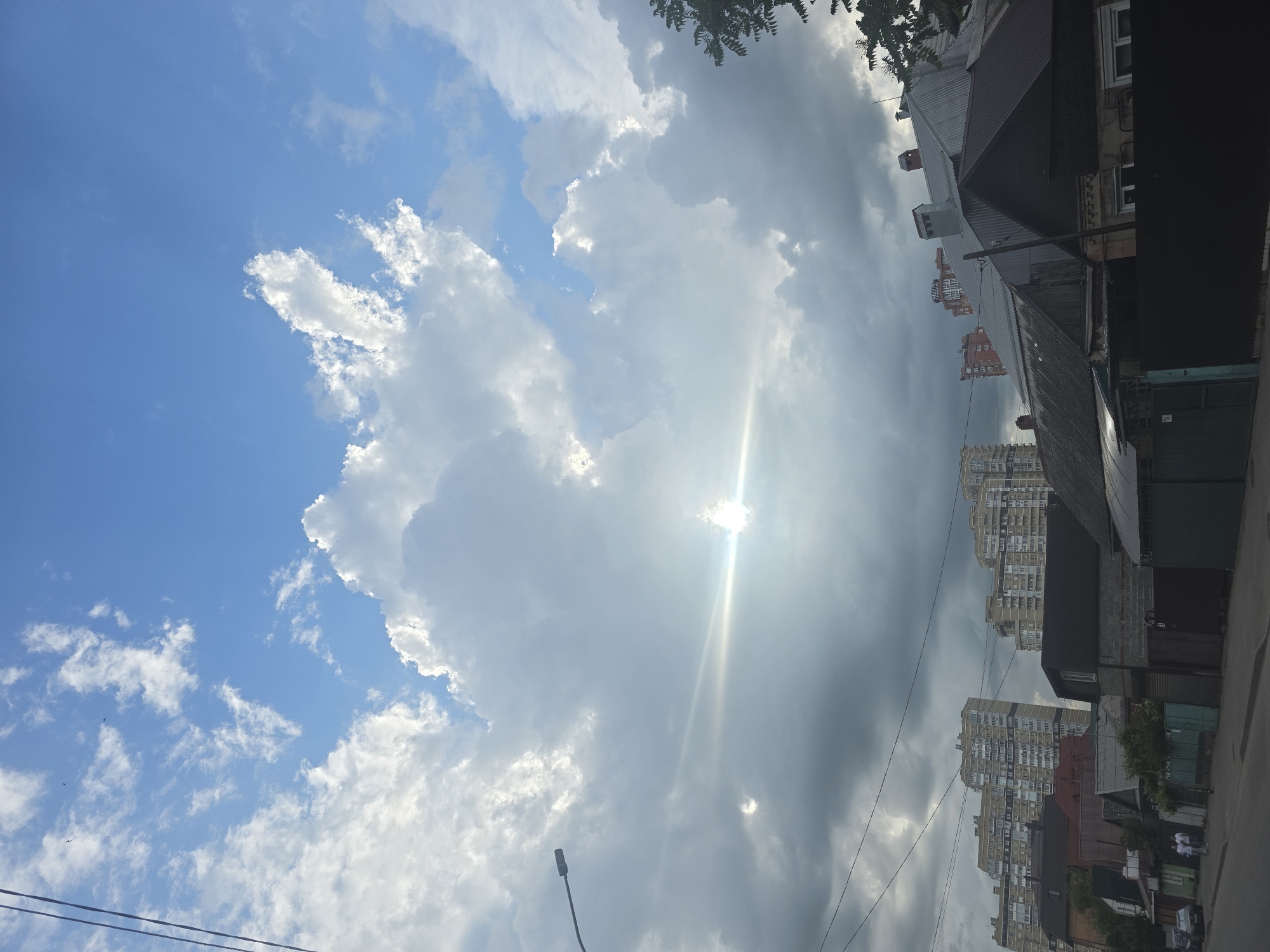 The image shows an upward view of a street scene in Krasnodar, Russia. The sky is partially cloudy with patches of blue sky visible. The sun is shining through the clouds, creating a bright glare. Buildings are visible on the right side of the frame, including residential apartments and smaller structures with roofs. Overhead power lines are strung between buildings. A streetlamp is visible in the lower left corner of the frame. The image captures a mix of urban and natural elements, typical of a city environment.
