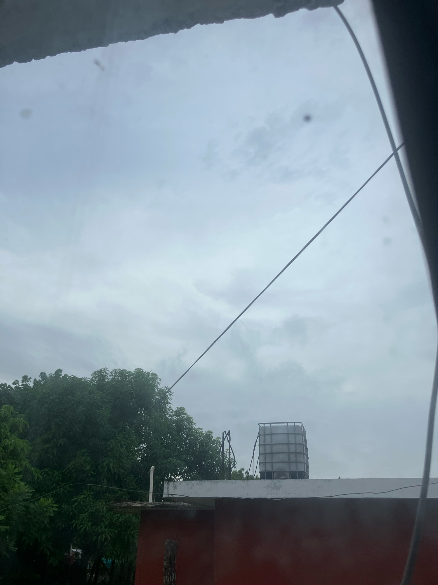 A cloudy sky with a large water tank on top of a building in Ébano, Mexico. The water tank is white and rectangular, with a metal cage around it. The building is red and white. There are trees in the background. The image is taken from a window, with a wire running across the frame.