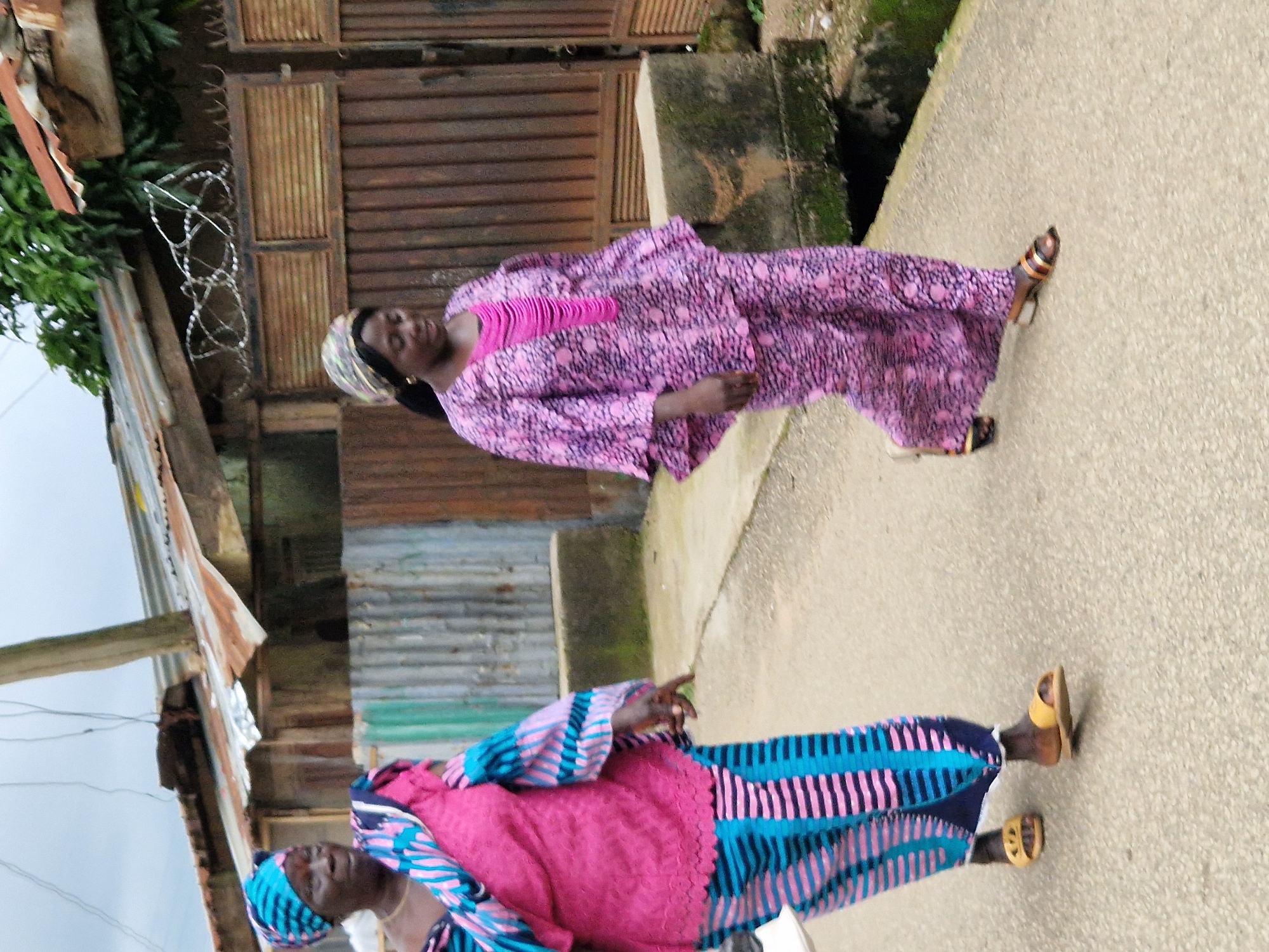 Two women are walking on a paved pathway in front of a weathered building with a corrugated metal roof and siding. The woman on the left is wearing a blue and pink headwrap, a pink top with lace detailing, and a blue and pink striped skirt. She is also wearing yellow sandals. The woman on the right is wearing a patterned headwrap, a long-sleeved purple patterned dress with a pink ruffled neckline, and sandals with gold and brown straps. The building behind them appears to be in disrepair, with rusted metal and exposed wood. There are trees and vegetation visible above the building. The sky is overcast.