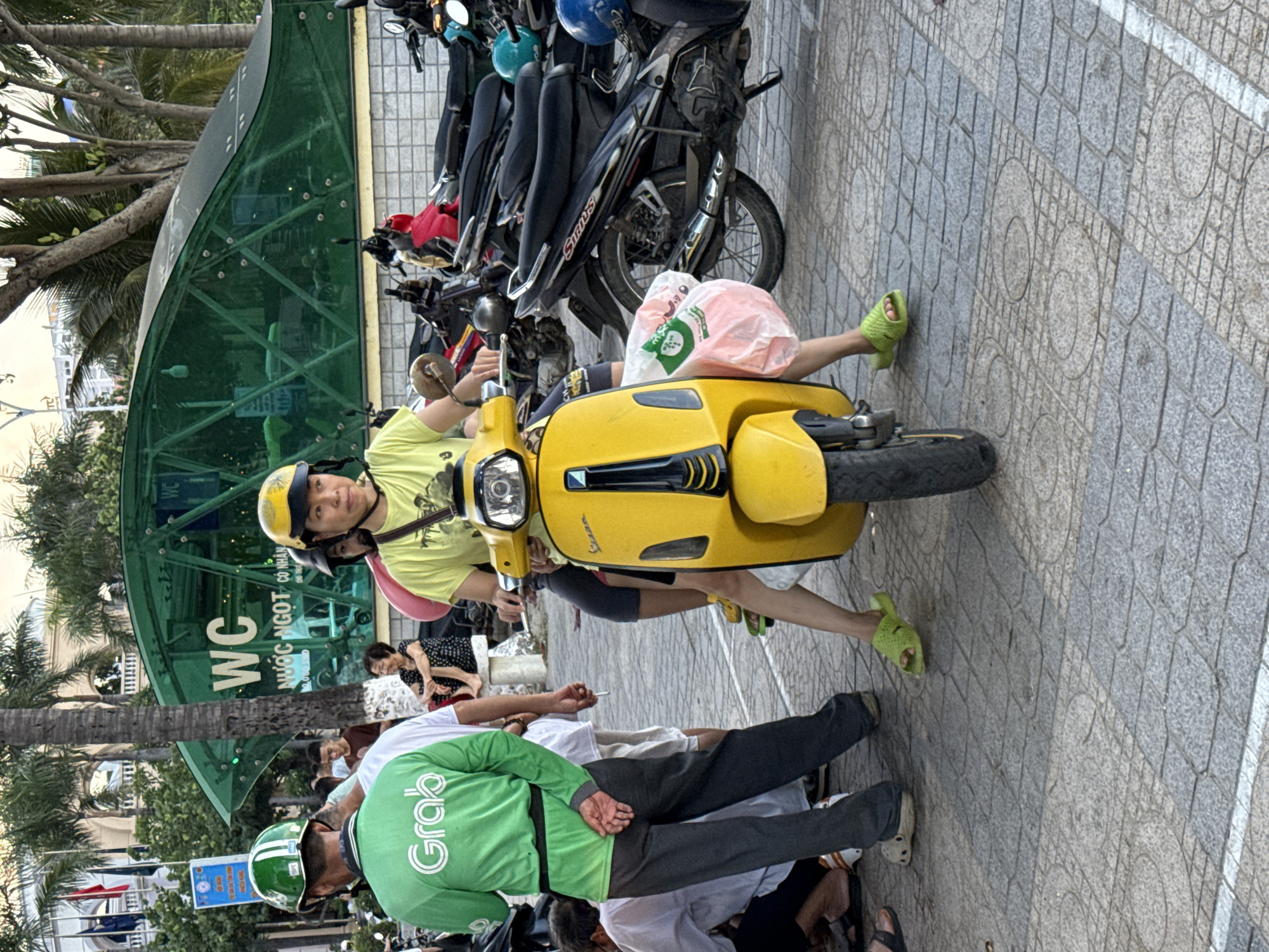 A group of people are gathered around parked motorcycles and a yellow scooter in an outdoor urban setting. In the foreground, a person wearing a bright yellow helmet and shirt is seated on the yellow scooter, holding a pink plastic bag. Another person, wearing a green long-sleeved shirt with the "Grab" logo, stands to the left, facing away from the camera. Several motorcycles are parked behind the scooter and in the background. A green, modern-looking building with "WC" signage is visible in the background. The ground is paved with square tiles. The overall scene suggests a public area in a city, possibly a marketplace or a transportation hub, in Nha Trang Ward, Vietnam.