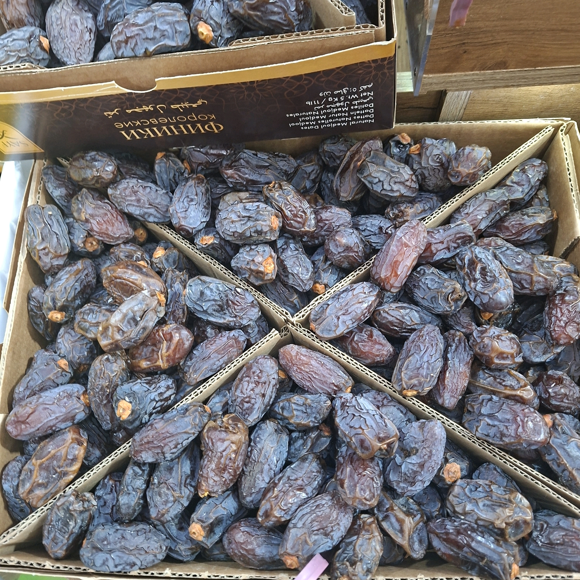 A close-up, overhead shot shows a display of dark, dried dates packed into cardboard boxes. The dates are wrinkled and have a slightly glossy sheen. Some of the dates have visible stems. The boxes are divided into four sections, with the dates filling each section. The background is blurred, but appears to be a market or retail setting with other boxes and wooden surfaces. Text on the side of one box is visible, containing Arabic script and English text that reads "Natural Medjool Dates." The scene is lit with natural or overhead lighting.