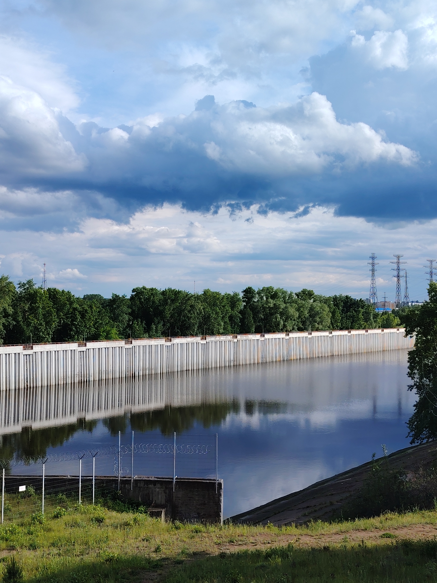 The image shows a waterfront view in Chaikovsky, Russia. A long, concrete flood wall stretches across the middle ground, reflecting in the water. Behind the wall, a line of green trees is visible, with power lines in the distance on the right side. The sky above is filled with dramatic, dark clouds. In the foreground, there is a grassy area with a fence and barbed wire. A concrete structure is visible behind the fence. The overall lighting suggests daylight, and the scene appears calm and still.