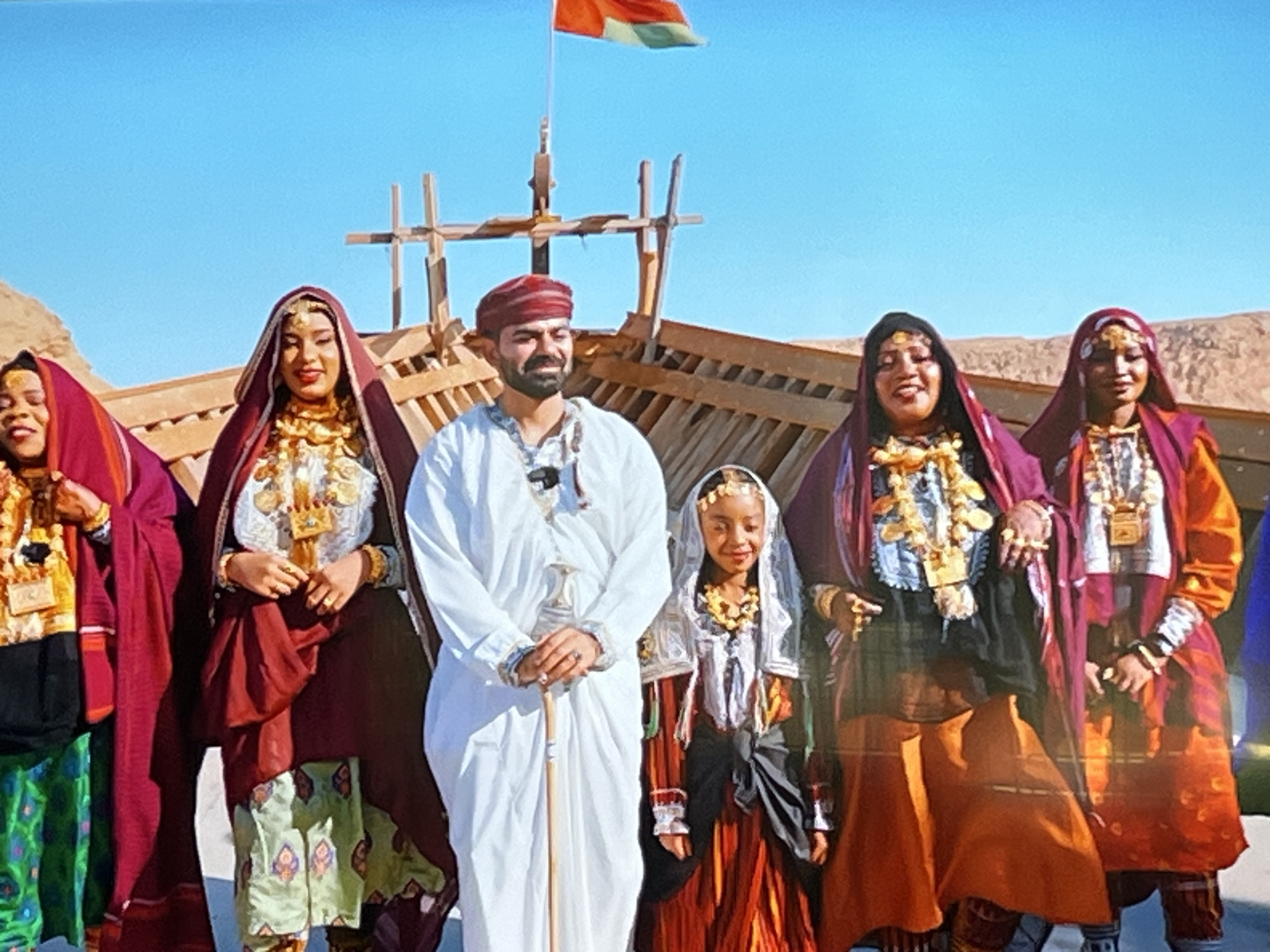 A group of six people, including one child, are posing for a photograph in front of a wooden ship structure against a clear blue sky. The individuals are dressed in traditional clothing adorned with gold jewelry. The person in the center, a man wearing a white robe and a red headscarf, holds a cane. The other five individuals, all women or girls, wear colorful robes, headscarves, and elaborate necklaces. The background shows a wooden ship skeleton, some desert-like terrain, and a flag flying from a pole. The image appears to be taken outdoors in a bright, sunny environment. There is no indication in the image that the location is Kharkiv, Ukraine.