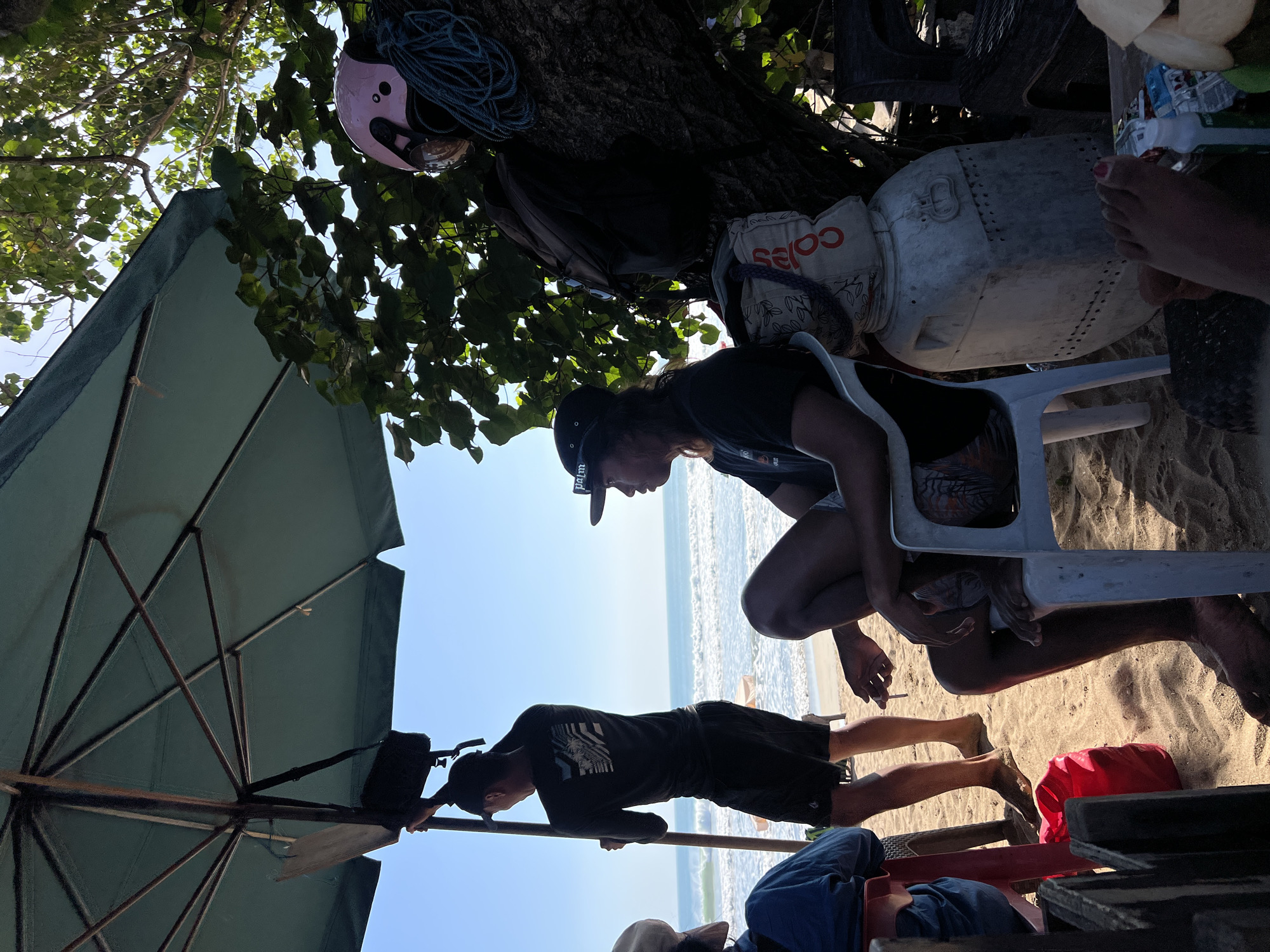 In Kuta, Indonesia, on a sandy beach, an umbrella provides shade for a group of people. A man stands holding a pole that supports the umbrella, wearing a black long-sleeved shirt and shorts. To his right, another man sits in a chair, also wearing a black t-shirt and shorts, and holding a cigarette. A woman's legs are visible in the foreground, one with painted toenails. The background shows the ocean with waves. A pink helmet and a coil of rope are visible in a tree. Several bags and a large plastic container are also present on the sand.