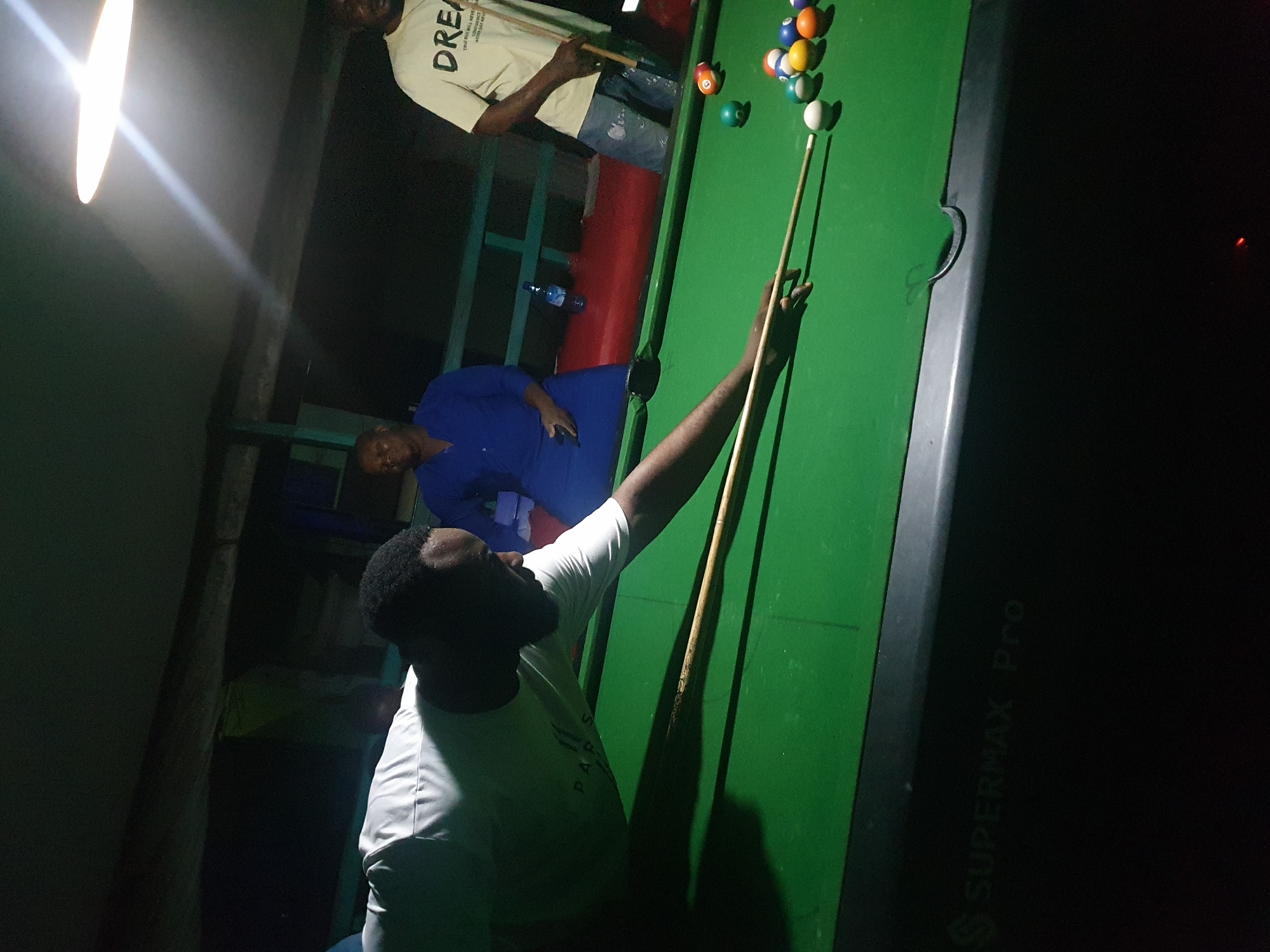 A group of men are playing pool in an indoor dimly lit space, likely a bar or recreational area. The table is green with several billiard balls arranged on it. One man in a white t-shirt is in the foreground, bent over the table and aiming a cue stick at a ball. Another man in a blue shirt is standing behind him, and a third man in a cream-colored t-shirt is further back, also holding a cue stick. The environment appears to be somewhat dated, with visible wooden beams and a fluorescent light fixture overhead. The text "SUPERMAX Pro" is visible on the side of the pool table. While the exact location in Lokoja, Nigeria, cannot be determined from the image alone, the general setting suggests a common recreational establishment.