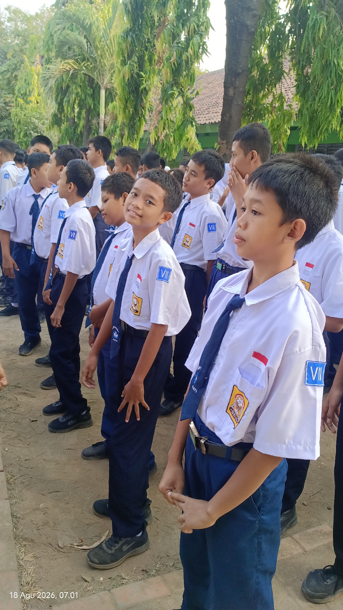 In Kecamatan Pati, Indonesia, a group of young boys in white shirts and navy blue pants stand in a line, some looking at the camera. The boy in the foreground, facing the camera, has a bright smile. He wears a white shirt with a navy blue tie and a blue patch on his sleeve with the Roman numeral "VII". Another boy to his left, also in uniform, is looking away from the camera with a neutral expression. The boys are standing on a dirt ground with some greenery and trees in the background, suggesting an outdoor school setting. The image is dated "18 Agu 2026, 07.01".