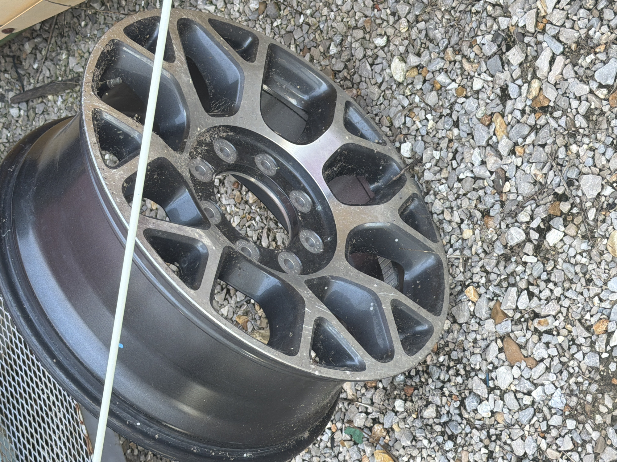 An aluminum alloy wheel, likely from a vehicle, rests on a gravel surface. The wheel is dark gray with silver accents, featuring a complex spoke design with circular cutouts. A light-colored flexible tube is draped across the face of the wheel. The gravel is composed of various small rocks and some dried vegetation, suggesting an outdoor environment. The scene is located in Hornersville, United States.