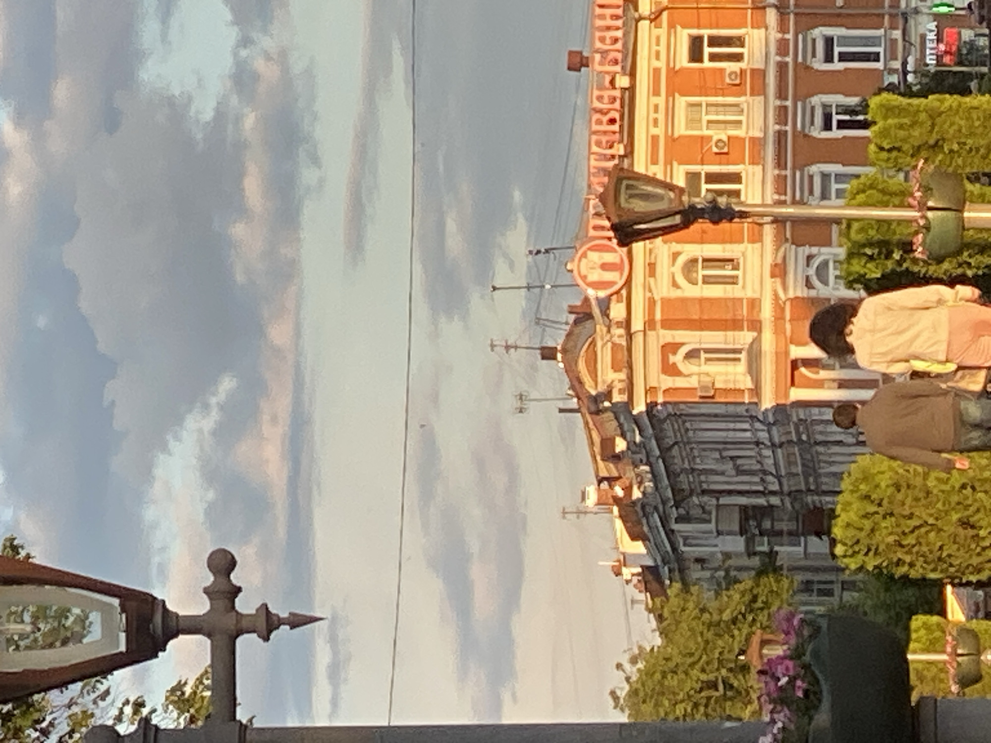 This image captures a street scene in an urban environment, likely a city center, under a partly cloudy sky. The photograph appears to be taken during the "golden hour," either early morning or late afternoon, given the warm light on the buildings and the hues in the sky.
In the foreground on the left, a decorative metal street lamp post is partially visible. The sky occupies a significant portion of the upper left and central parts of the image, displaying a mix of light blue, grey, and subtle pinkish clouds.
On the right side of the image, two distinct buildings dominate the cityscape. The most prominent building, an orange-ochre structure with white window frames and architectural detailing, stretches across the mid-ground. Several air conditioning units are visible beneath the windows. Along the top of this building, Cyrillic text is clearly legible, reading "ЦЕНТРАЛЬНИЙ УНІВЕРМАГ" (Tsentralnyy Univermah), identifying it as a "Central Department Store." Further to the right, a smaller sign with "АПТЕКА" (Apteka), meaning "Pharmacy," is visible above some greenery. Behind the orange building and to its left, a darker, possibly grey or blue, multi-story building with numerous windows is visible.
In the lower right, two individuals are partially visible. One person, wearing a light-colored top, stands near a green bush, while another person in a brown jacket and blue jeans is partially obscured by dense green foliage, possibly sitting or bending down. A street light or traffic light pole is also present in this area, featuring a red circular sign with a white capital "H." Ornamental green bushes or hedges line the foreground and mid-ground on the right.
The overall scene depicts a bustling, well-maintained city street, consistent with a commercial district, located in Poltava, Ukraine, as indicated by the Cyrillic text.