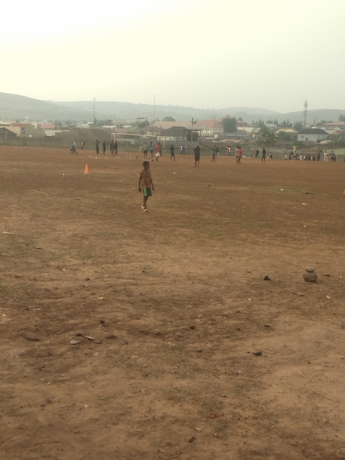 The image shows an open, dirt field in New Karu, Nigeria, at the coordinates 8.979 latitude and 7.565 longitude. The field appears to be used for playing soccer as there are several people playing and there is a small orange cone in the foreground. There is a single boy walking towards the camera in the center of the field. Several other people are playing soccer in the distance, and there is a low-lying hill in the background. The scene is overcast and a small amount of greenery can be seen in the field, along with some trash. There are several houses and buildings in the distance, indicating a residential area.
