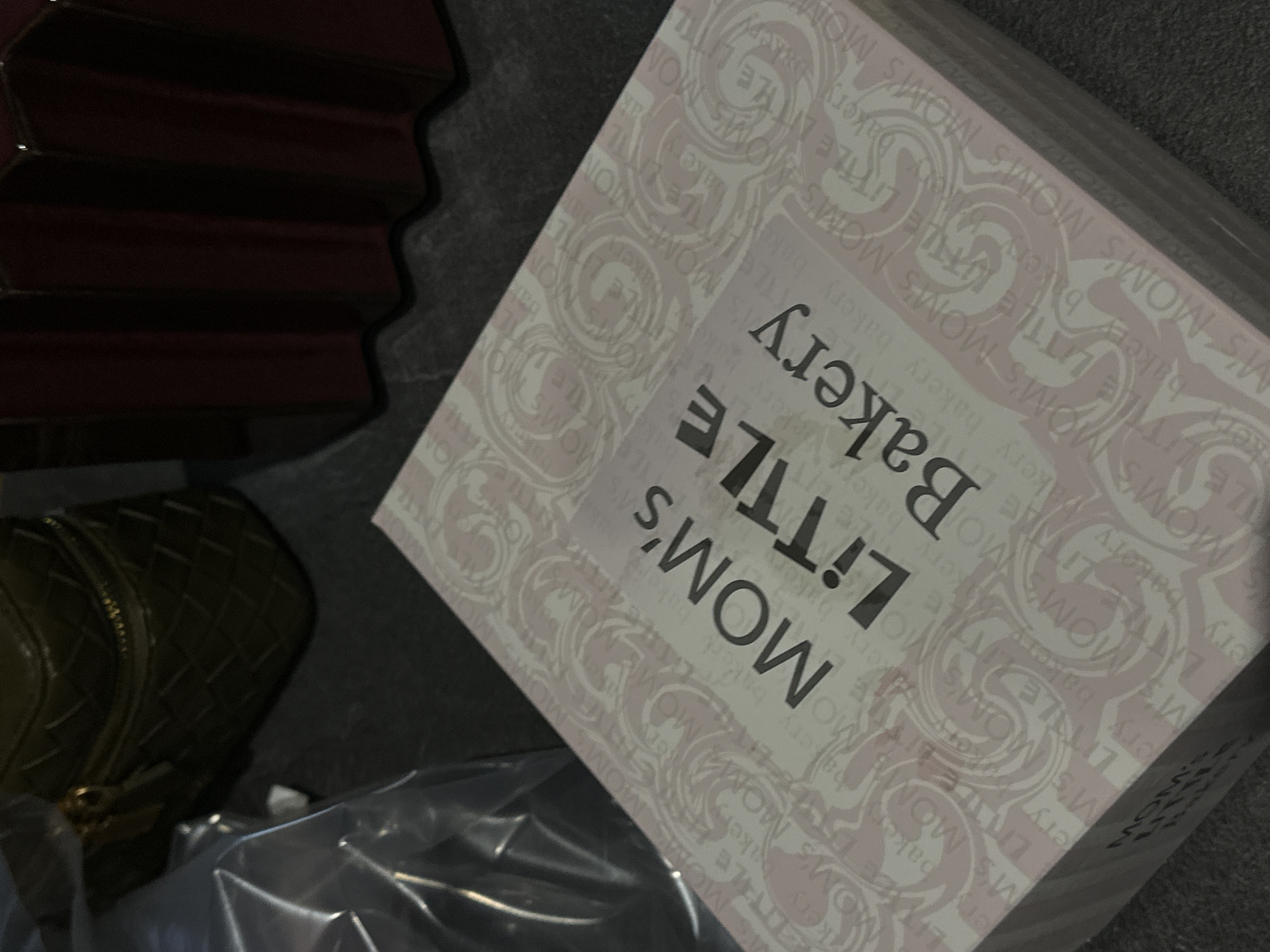The image shows a white box from Mom's Little Bakery, situated next to a dark green, quilted purse and some clear plastic wrapping. The box is decorated with a pink and white floral pattern and the bakery's name is printed in black. The location is most likely in Yerevan, Armenia, given the bakery's name. A dark object is visible in the top left corner, which appears to be a piece of furniture. The surface the objects are resting on is grey.