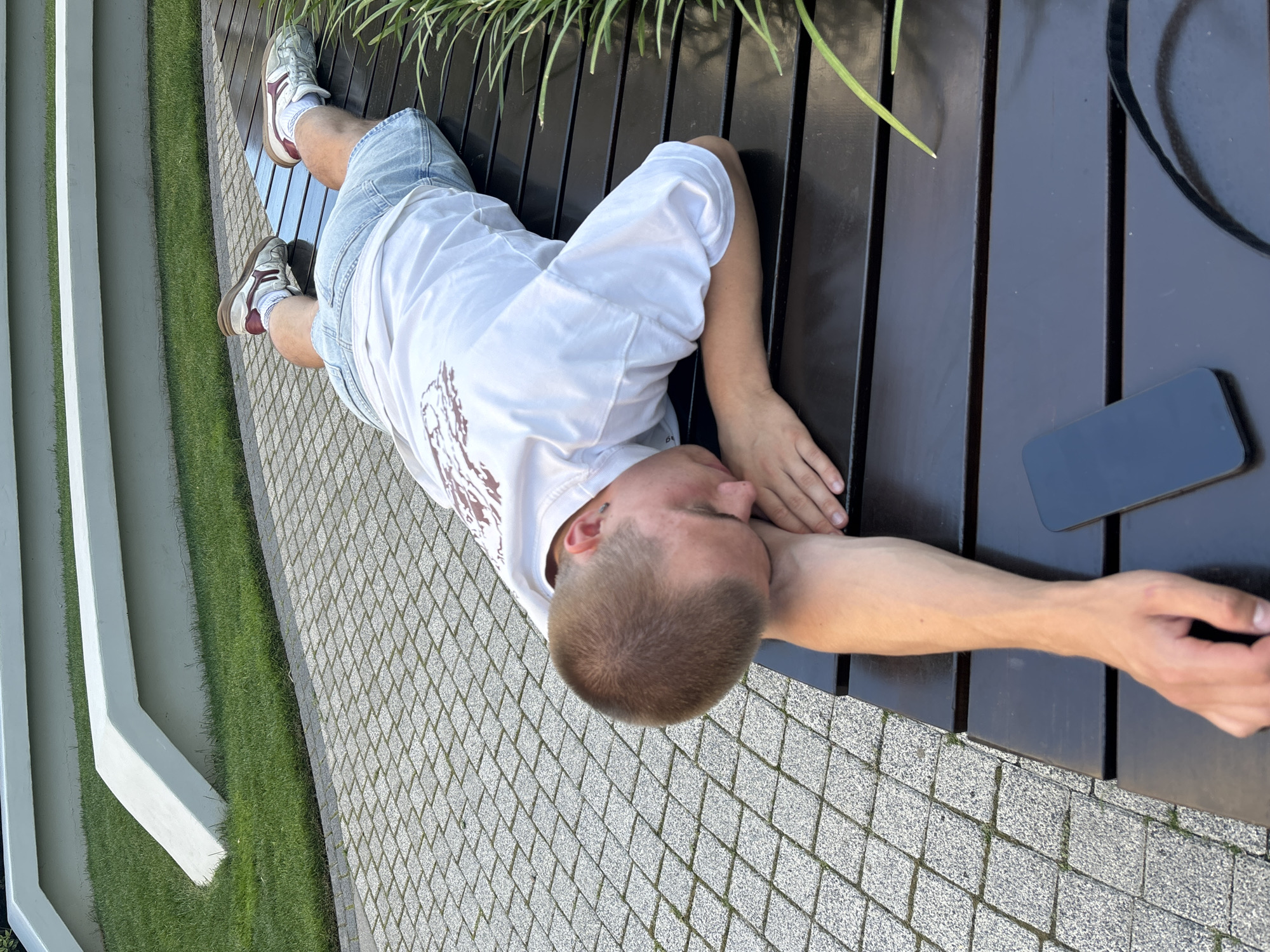 A young person with short brown hair lies face down on a dark wooden bench with their head resting on their bent arm. They are wearing a white t-shirt with a graphic design and light blue denim shorts. Their feet, clad in white socks and athletic shoes with maroon accents, are visible at the top of the frame. A black smartphone lies on the bench near their legs. The bench is situated on a patterned stone walkway, with a strip of green grass and white concrete structures in the background. The setting appears to be an outdoor public space, possibly a park or plaza, in Belgrade, Serbia.
