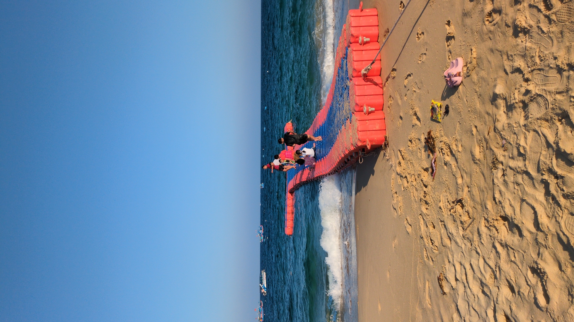 The scene depicts a sunny beach environment in East Quy Nhon Ward, Vietnam. A modular floating pathway, constructed from red plastic pontoons with a blue top surface, extends from the sandy beach into the blue-green ocean. The pathway forms a gentle ramp downwards, with a small T-section at its seaward end.
Approximately five to seven individuals are present on the floating pathway. One person, wearing a black shirt and dark shorts, stands barefoot near the end of the pathway, facing the open sea. Another individual, dressed in a white shirt and dark bottoms, is seated on the pathway closer to the shore. Other individuals are further along the pathway, some standing, some seated. One person in a red shirt is partially visible.
Ocean waves are breaking against the floating pathway where it meets the water. In the distance, numerous boats are visible on the horizon, including several traditional Vietnamese fishing vessels and at least one white speedboat.
The foreground consists of light brown sand, marked with numerous footprints. A pair of light pink sandals, a yellow wrapper, and natural debris such as leaves and twigs are visible on the sand near the pathway's entry point. A dark rope or cable anchors the floating pathway to the sand in the lower right corner. The sky above is clear, cloudless, and a uniform light blue.