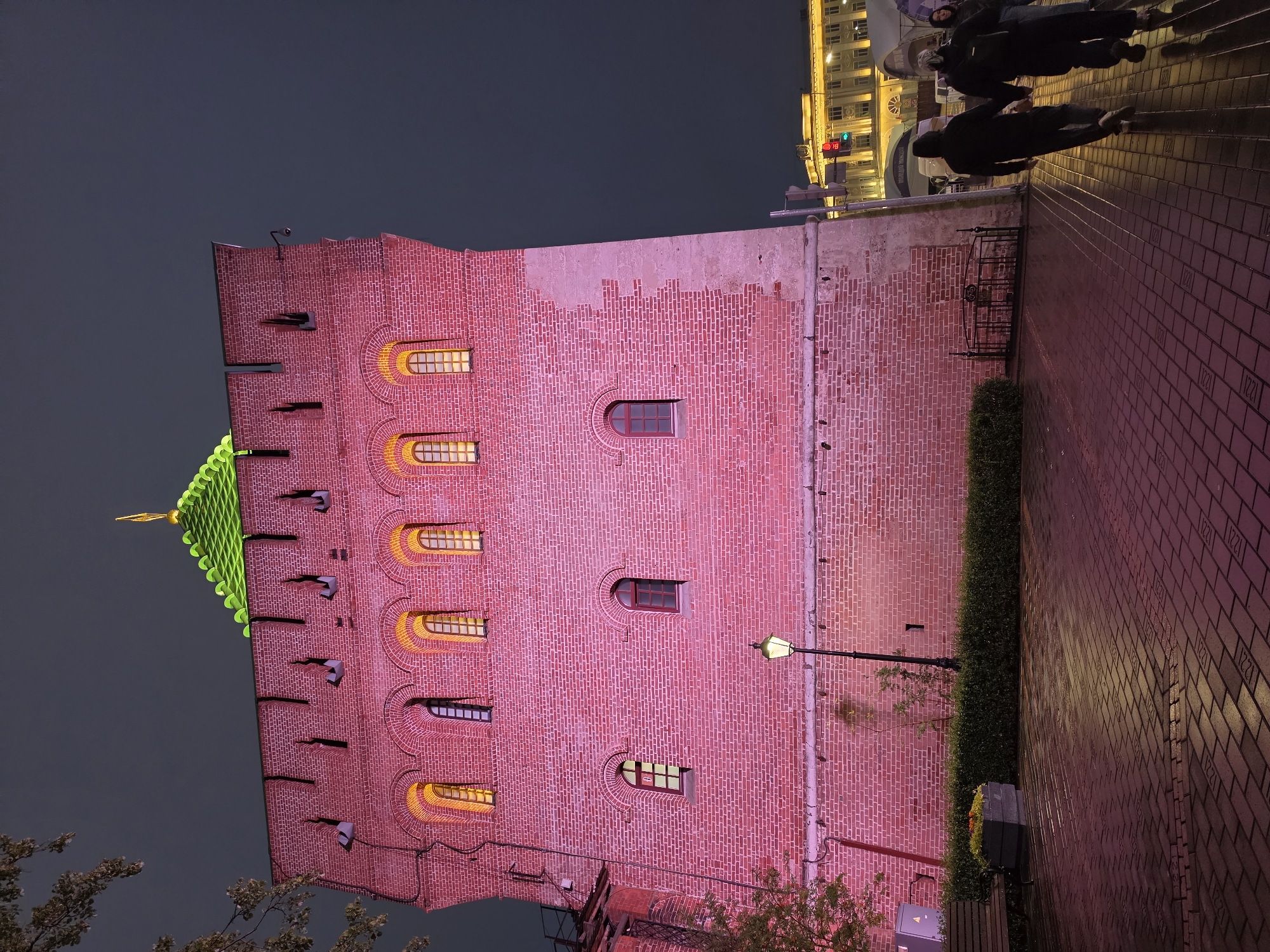 A nighttime, eye-level shot captures the red brick tower of the Nizhny Novgorod Kremlin, illuminated by pink and yellow lights. The tower's crenellated top and green roof are visible against a dark sky. Several arched windows with yellow light emanate from the upper floors, while smaller, dark windows are present on the lower levels. A single lamppost stands near the base of the tower.
In the foreground, a paved walkway with a patterned brick surface reflects the pink light. A low green hedge runs along the base of the tower, with a planter box and a bench visible to the left. To the right of the tower, a couple walks away from the camera, hand-in-hand, their figures silhouetted by the ambient lighting. Behind them, a street scene unfolds with illuminated buildings, traffic lights showing red and green, and a few other pedestrians. The overall impression is of an ancient structure bathed in modern light in an urban setting.