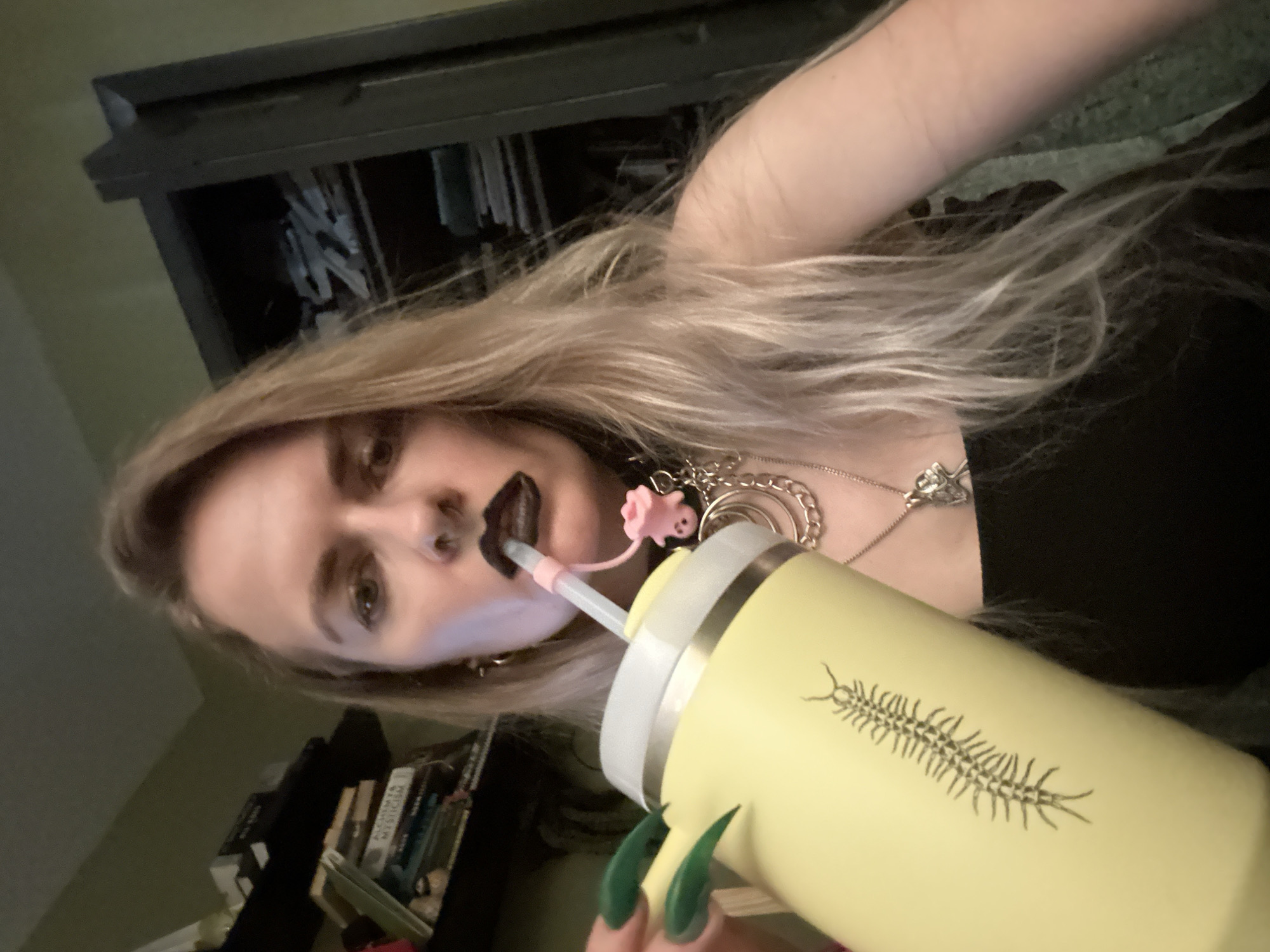 A selfie-style photograph shows a blonde-haired woman drinking from a light yellow tumbler with a white lid and a drawing of a centipede on it. A straw extends from the tumbler to the woman's mouth, which is painted with dark lipstick. She has several necklaces and long, green fingernails. The background shows a cabinet and a bookshelf with books. The location in Cape Coral, United States of America is not discernible from the image.
