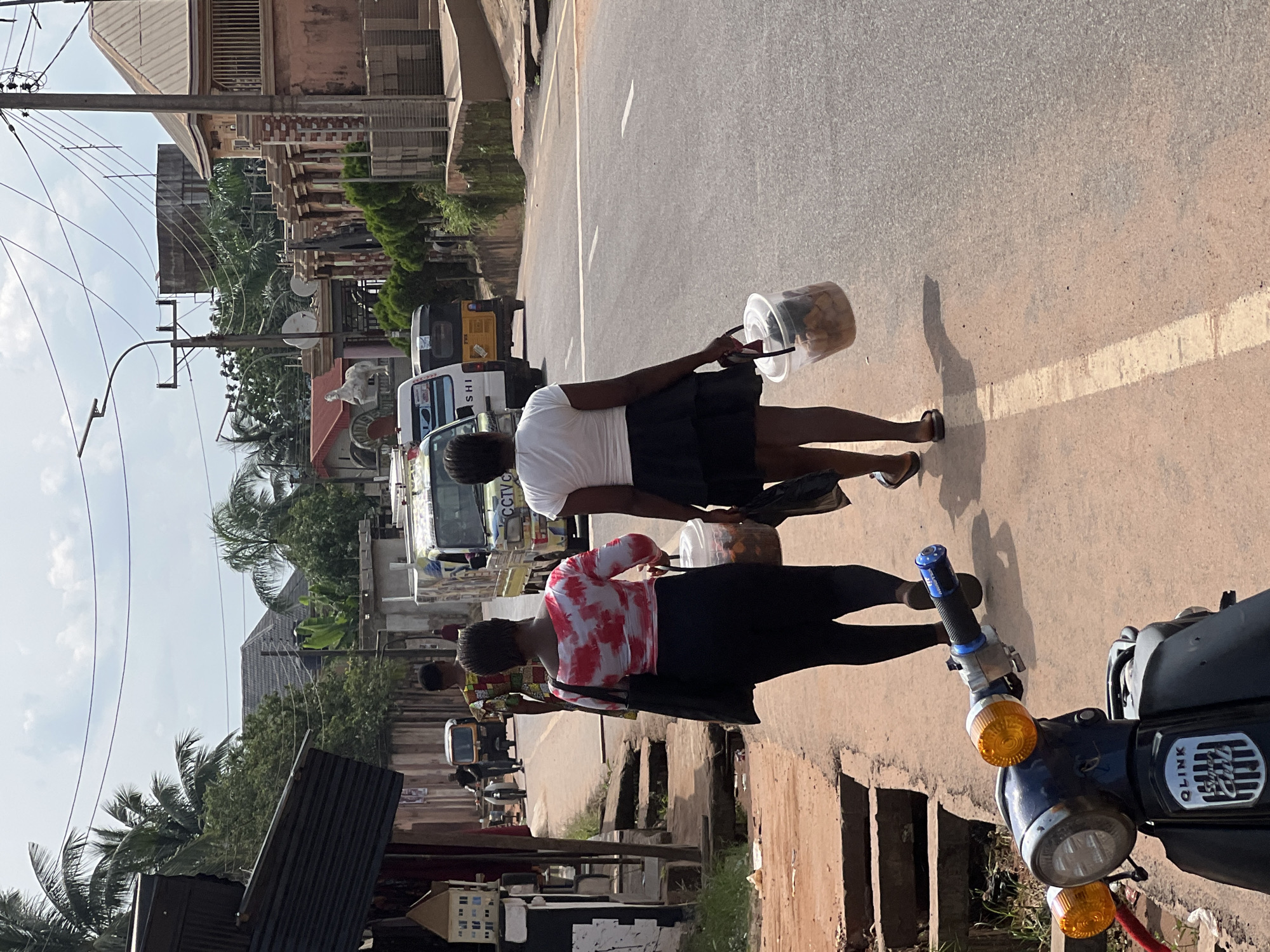 Two young women walk along a paved road in Nkpor, Nigeria. The woman in the foreground on the left wears a red and white tie-dye off-the-shoulder top and black leggings, carrying a black shoulder bag and a clear plastic bucket filled with food. The woman on the right wears a white off-the-shoulder top and a black skirt, also carrying a clear plastic bucket filled with food. They are walking away from the viewer on a sidewalk lined with concrete drainage channels. In the background, vehicles including a yellow tricycle, a white van with "CCIVC" lettering, and a motorcycle are visible on the road. The environment appears to be a moderately developed urban area with buildings and vegetation lining the street. The sky is partly cloudy.