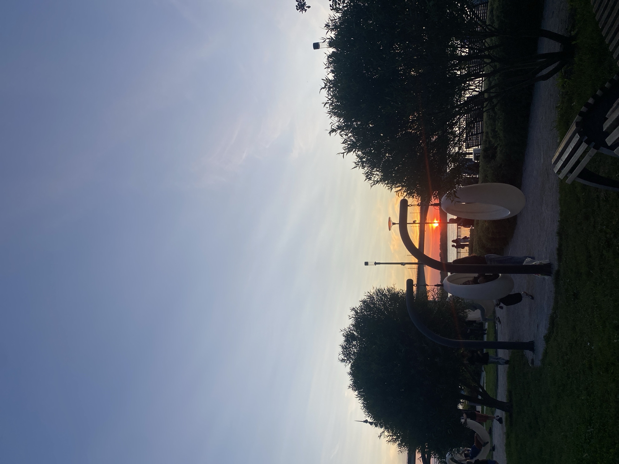 This is a medium-shot, eye-level photograph of a sunset over the Kama River in Perm, Russia. The sky is a gradient of soft blues and pale oranges, with a few wispy clouds. The sun, a bright orange orb, is low on the horizon, casting a warm glow. In the foreground, a park area is visible with trees, grass, and benches. Two modern, circular swings hang from curved metal structures. Several people are scattered throughout the park, some sitting on benches, others walking or interacting with the swings. A bridge is visible in the distance, spanning the river. The overall atmosphere is peaceful and serene.