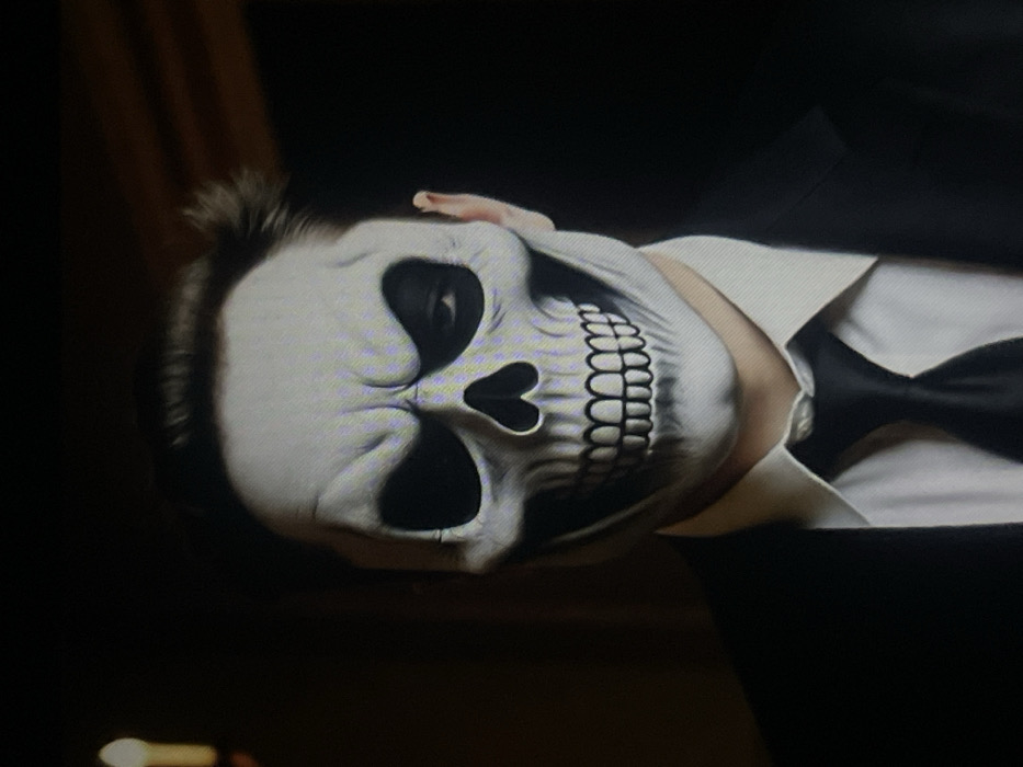 A single individual is depicted in a medium close-up, standing in a dimly lit indoor setting in Muchiya, Nigeria. The subject, presumed male, is wearing a black suit jacket, a white collared shirt, and a black necktie. A white and black skull-patterned mask covers the entirety of the subject's face. The mask features prominent black eye sockets, a black nasal cavity, and a wide, stylized grin with delineated teeth. The subject's right eye is partially visible through the mask's eye opening. Dark hair is visible above the mask, protruding from the forehead area. A blurred, vertical light source, possibly a candle or a small lamp, emits a soft yellow-orange glow in the upper left background. The background is otherwise dark and indistinct. The subject is stationary, facing directly towards the viewer.