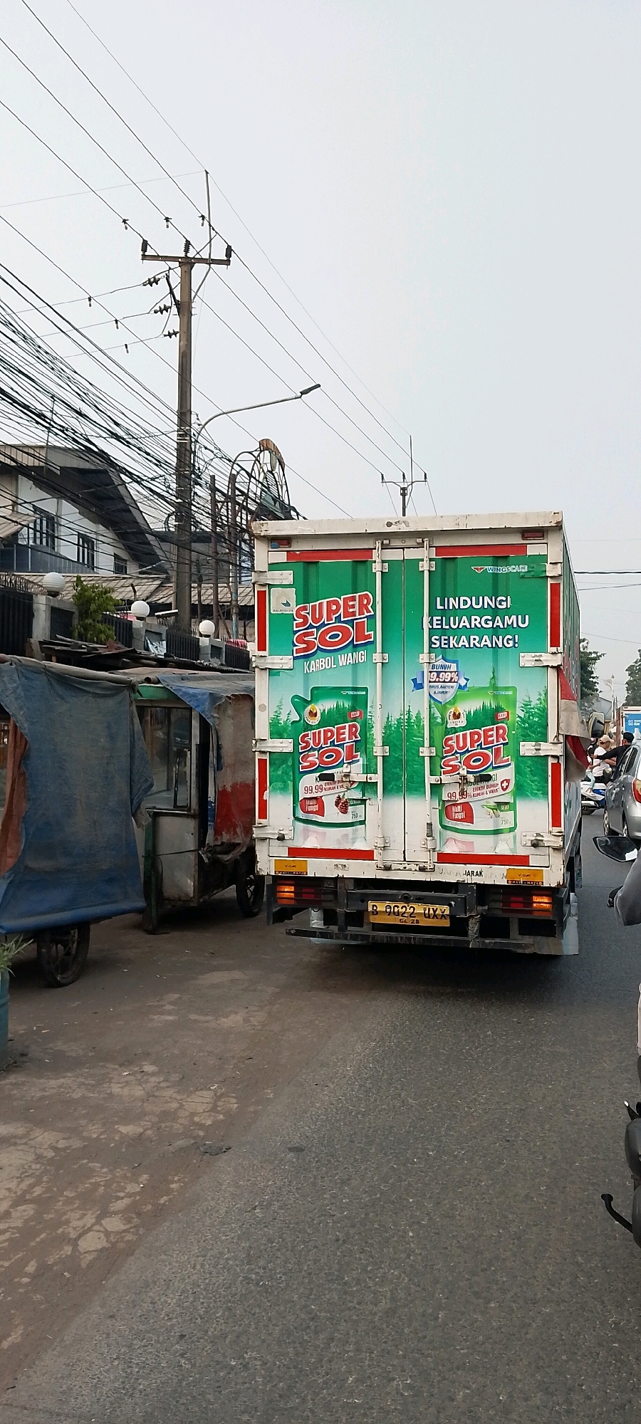 The image captures a street scene in Cileungsi, Indonesia, focusing on the rear of a white truck with green branding. The truck is advertising "SUPER SOL" disinfectant, with prominent text stating "LINDUNGI KELUARGAMU SEKARANG!" (Protect your family now!). The rear doors of the truck display two identical advertisements for "SUPER SOL" featuring images of bottles, pine trees, and the text "99.99% MULUS, BAKTERI & JAMUR" (99.99% Smooth, Bacteria & Fungi). A license plate, B 9022 UXX GC 2B, is visible on the rear of the truck.
To the left of the truck, a small, dilapidated food cart or stall is parked. It has a blue and red tarp covering its structure, and a single wheel is visible. The cart appears to be made of metal and wood.
The environment includes a paved road, likely asphalt, with some cracks and debris. The sidewalk area to the left is unpaved and rough. Overhead, a complex network of electrical wires and a wooden utility pole dominate the upper portion of the image. In the background, buildings with multiple stories and visible windows are present, along with a partial view of what appears to be a Ferris wheel-like structure. The sky is overcast and a pale gray. Other vehicles, including motorcycles and a car, are partially visible on the road to the right, suggesting traffic. No individuals are clearly visible or identifiable in the scene.