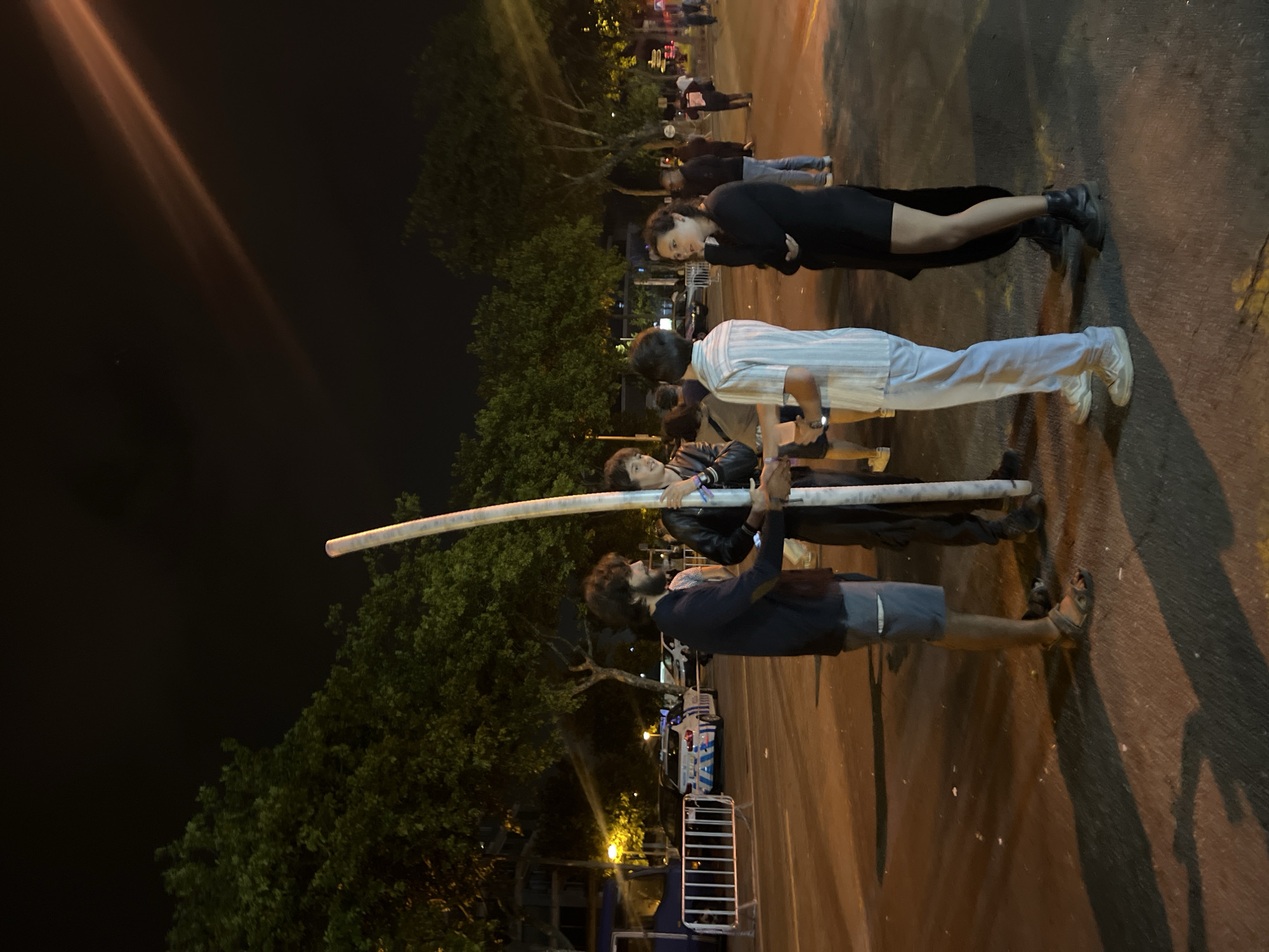 Four individuals are gathered outdoors at night, near a street with trees lining the background. The main focus is on a tall, cylindrical object, possibly a pole or a large pipe, held vertically by two men in the foreground. One man, with dark hair and a beard, wears a dark long-sleeved shirt and shorts, and sandals. He is shaking hands with another man, who is wearing a black leather jacket over a lighter shirt and dark pants. To the right, a third person in a striped shirt and light-colored pants is also present, facing the two men. A fourth individual, a woman in a black dress and boots, stands to the far right, looking towards the group. In the background, a police car with illuminated blue and white markings is visible, as well as metal barriers and more people in the distance. The scene appears to be in a public space, possibly a street or plaza, with streetlights casting warm illumination and creating long shadows on the asphalt. The location is identified as Matosinhos, Portugal.