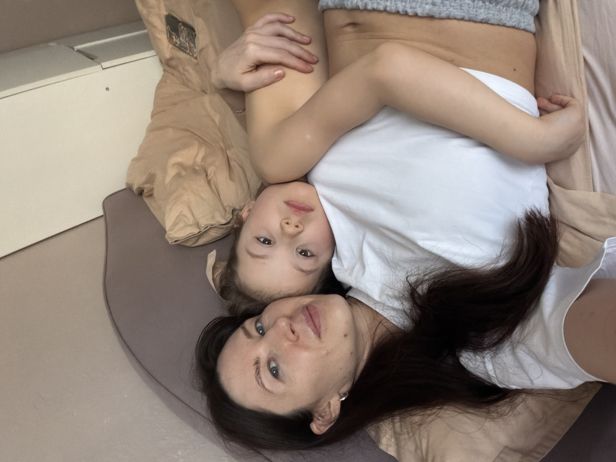 The image depicts an adult female and a young child lying together in an indoor domestic setting. The adult, with long dark hair, light skin, and blue eyes, appears to be in her 30s and is wearing a white short-sleeved top. She has a silver hoop earring in her visible left ear. Her left arm is wrapped around the child's torso. The child, a young girl with light brown hair and brown eyes, appears to be between three and seven years old. She is lying on her left side, resting her head on the adult's chest/shoulder area. She appears shirtless from the waist up, with grey knitted shorts visible on her lower body. Both individuals are looking directly at the camera. They are positioned on a bed or couch covered with light brown bedding. A purple/mauve headboard or pillow is visible behind the adult. In the upper right corner, a mobile phone with a patterned case is resting on the bedding, and a white wardrobe or cabinet is partially visible in the background. The specific location within the city of Perm, Russia, cannot be determined from the visual information provided.