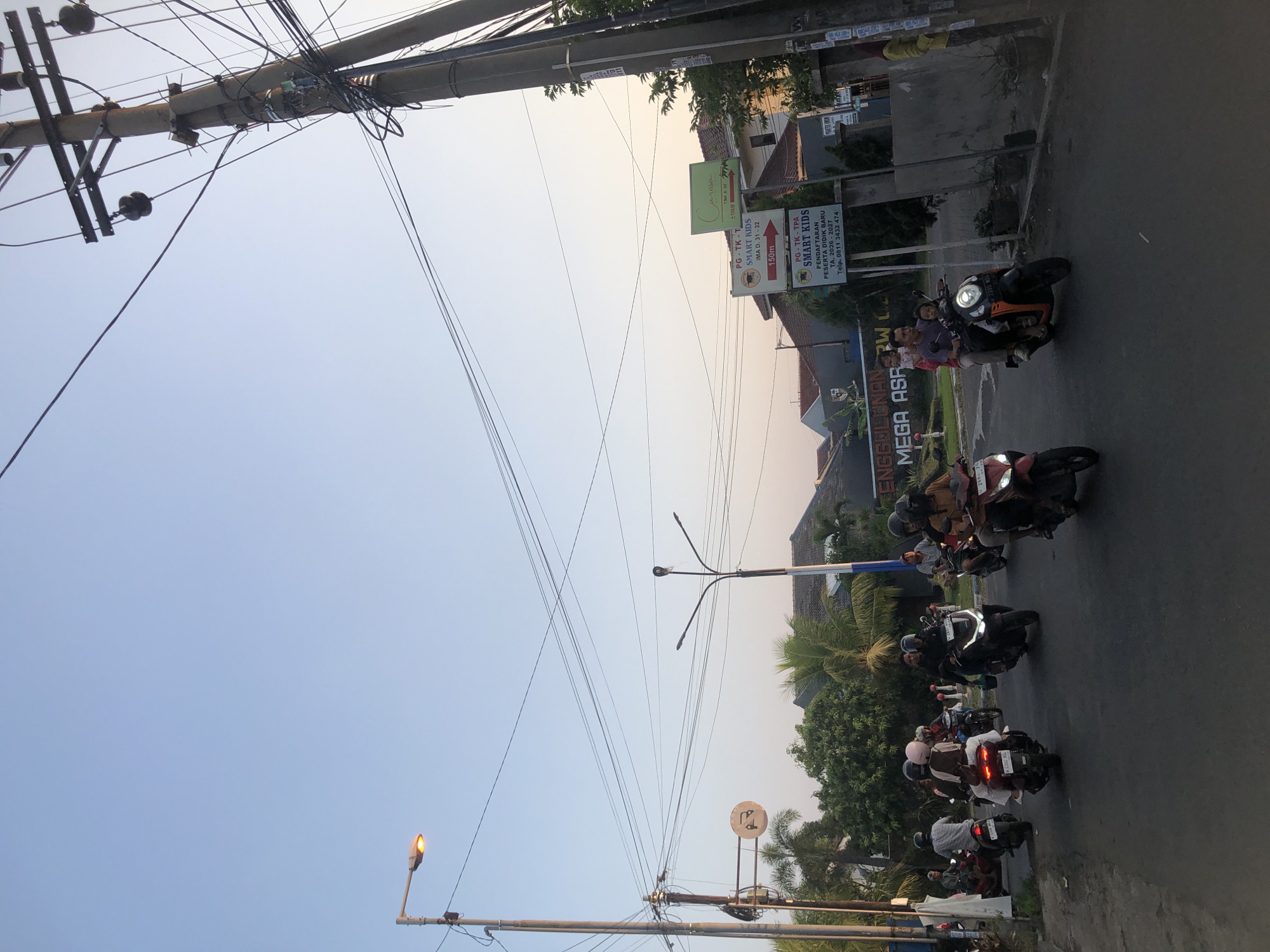 A street scene in Tenggulunan, Indonesia, features a group of people riding motorcycles on a paved road under a clear sky. A street light on the left side of the image is illuminated. Utility poles with numerous wires are visible throughout the scene, with one prominent pole on the right. Several motorcycles are in motion, with riders and passengers. Some buildings and trees are present in the background. Signs for "SMART KIDS" and "MEGA ASR" are visible on poles. The lighting suggests it is either early morning or late afternoon.