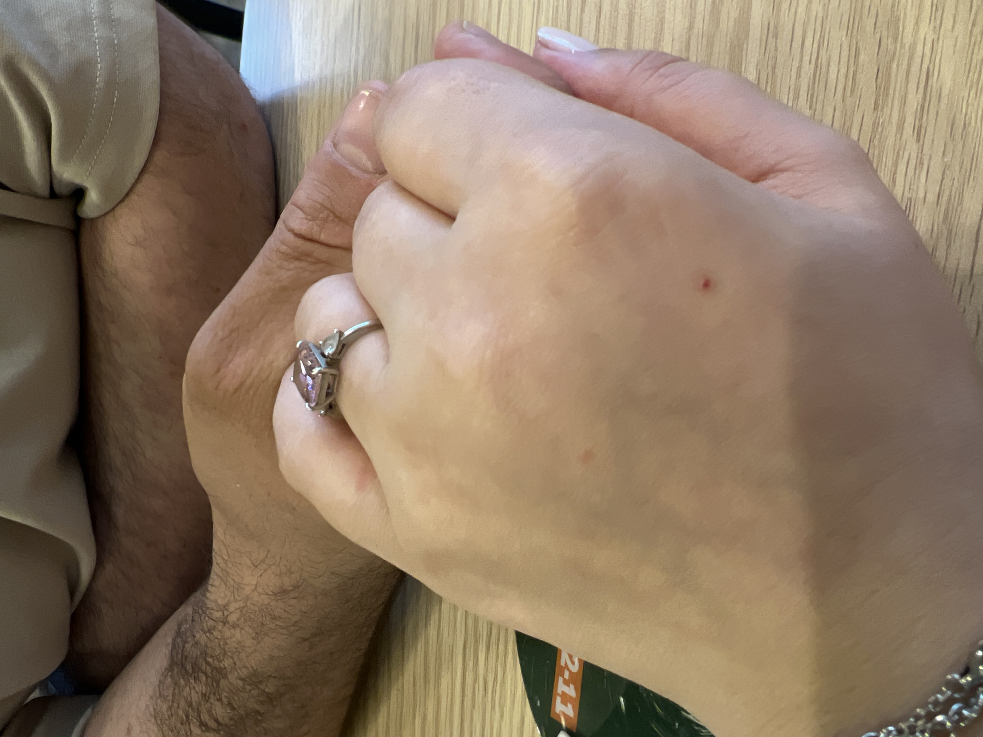 The media file is a close-up, overhead shot of two people holding hands. One person's hand, on the right, is lighter skinned and has a pink gemstone ring on their ring finger. The other person's hand, on the left, is darker skinned and appears to be covered in hair, suggesting it belongs to a man. The individuals are seated, as indicated by the visible parts of their legs, with one person wearing beige pants. They are holding hands over a light-colored, wooden surface, possibly a table. The lighting suggests it is daytime. There are no discernible weather cues. The scene evokes a sense of intimacy or connection between the two individuals. In the bottom right corner, a sliver of a green object with white and red text, partially obscured, is visible. The text includes "2-11". The location context is Yekaterinburg, Russia.